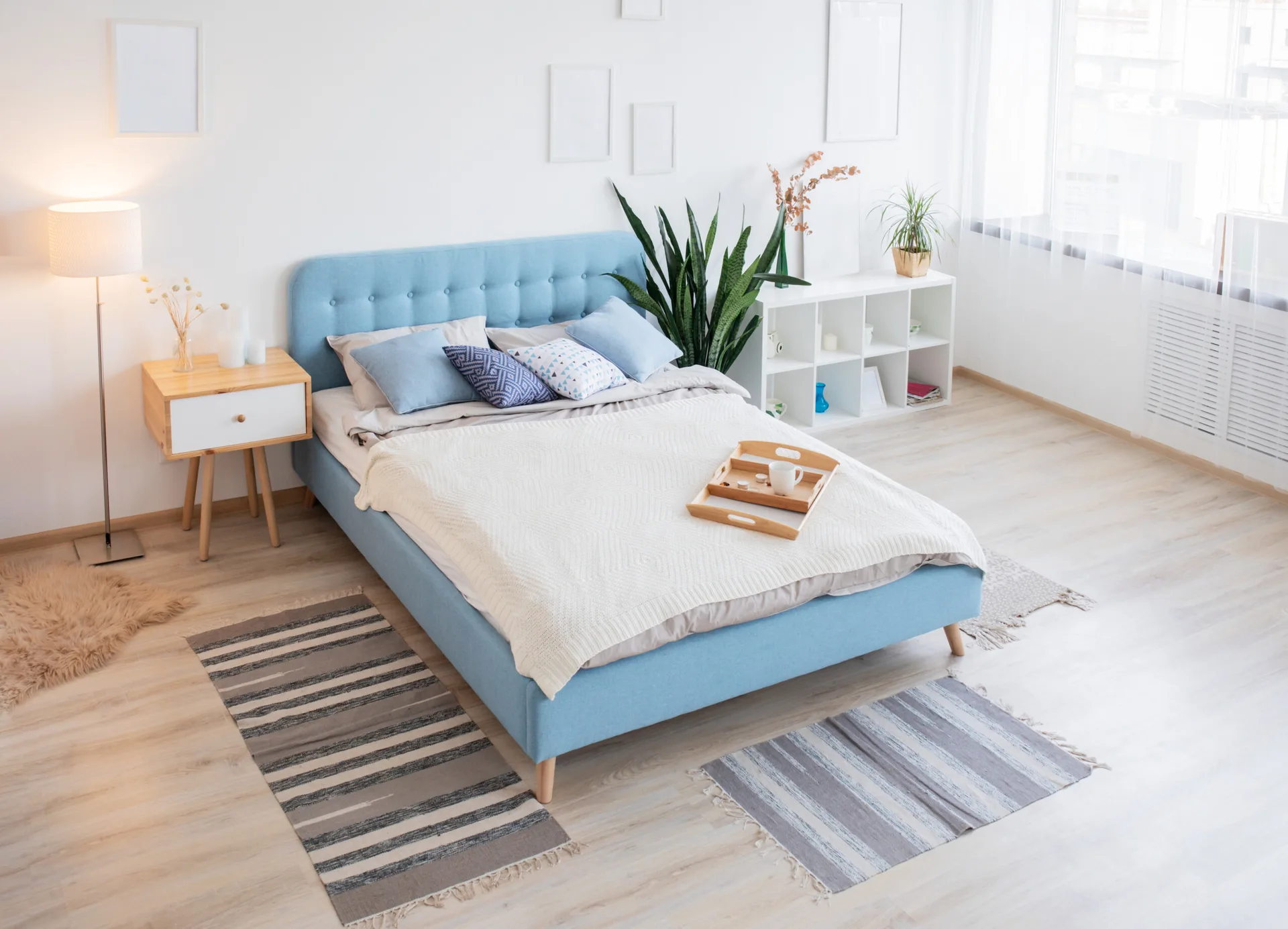 A bedroom features a blue tufted upholstered bed, light wood floors, striped accent rugs, and a wooden nightstand against white walls.