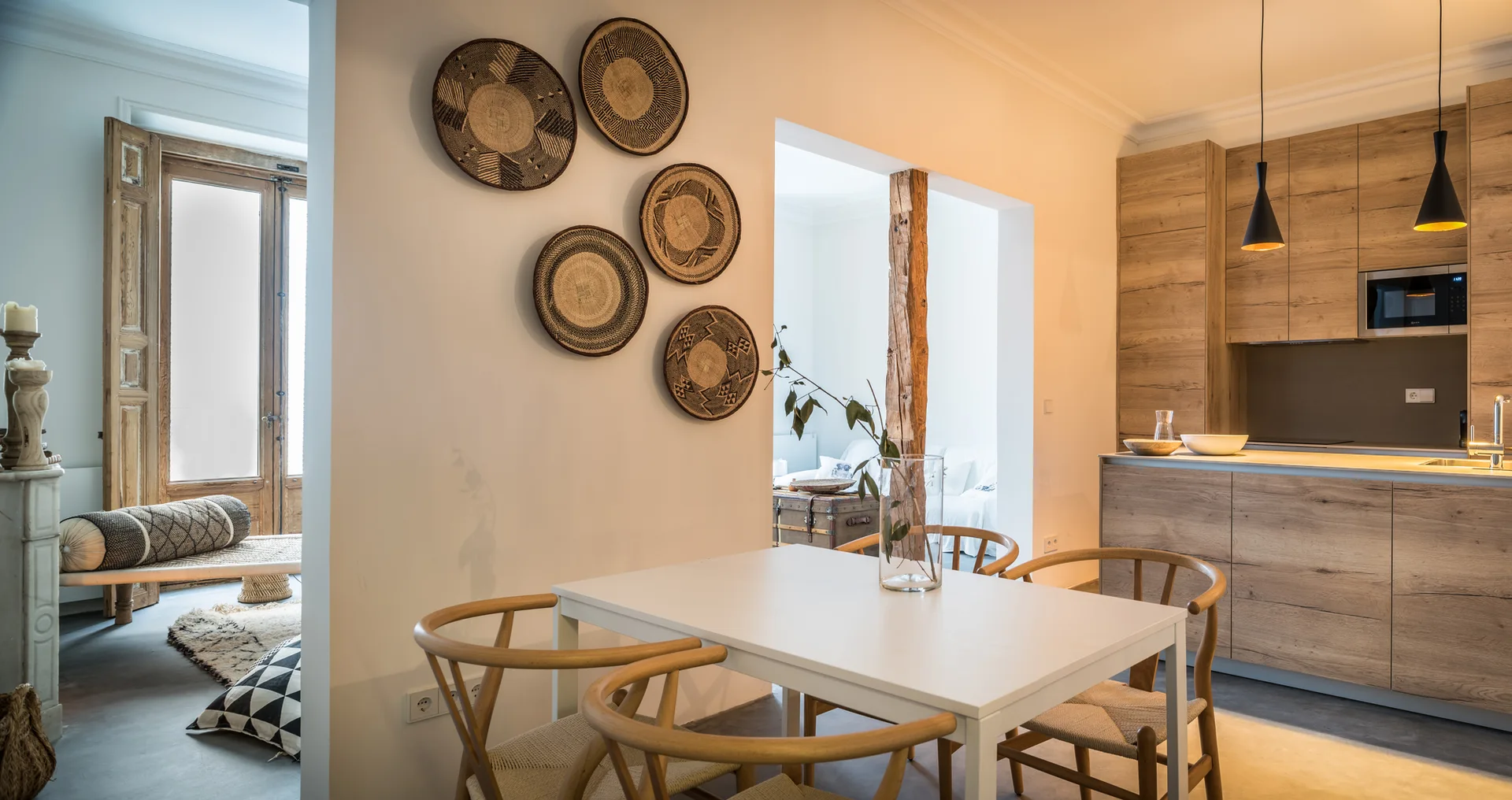 A white dining table with light wood chairs sits in front of a white wall decorated with five woven baskets, next to a wood-toned kitchen.