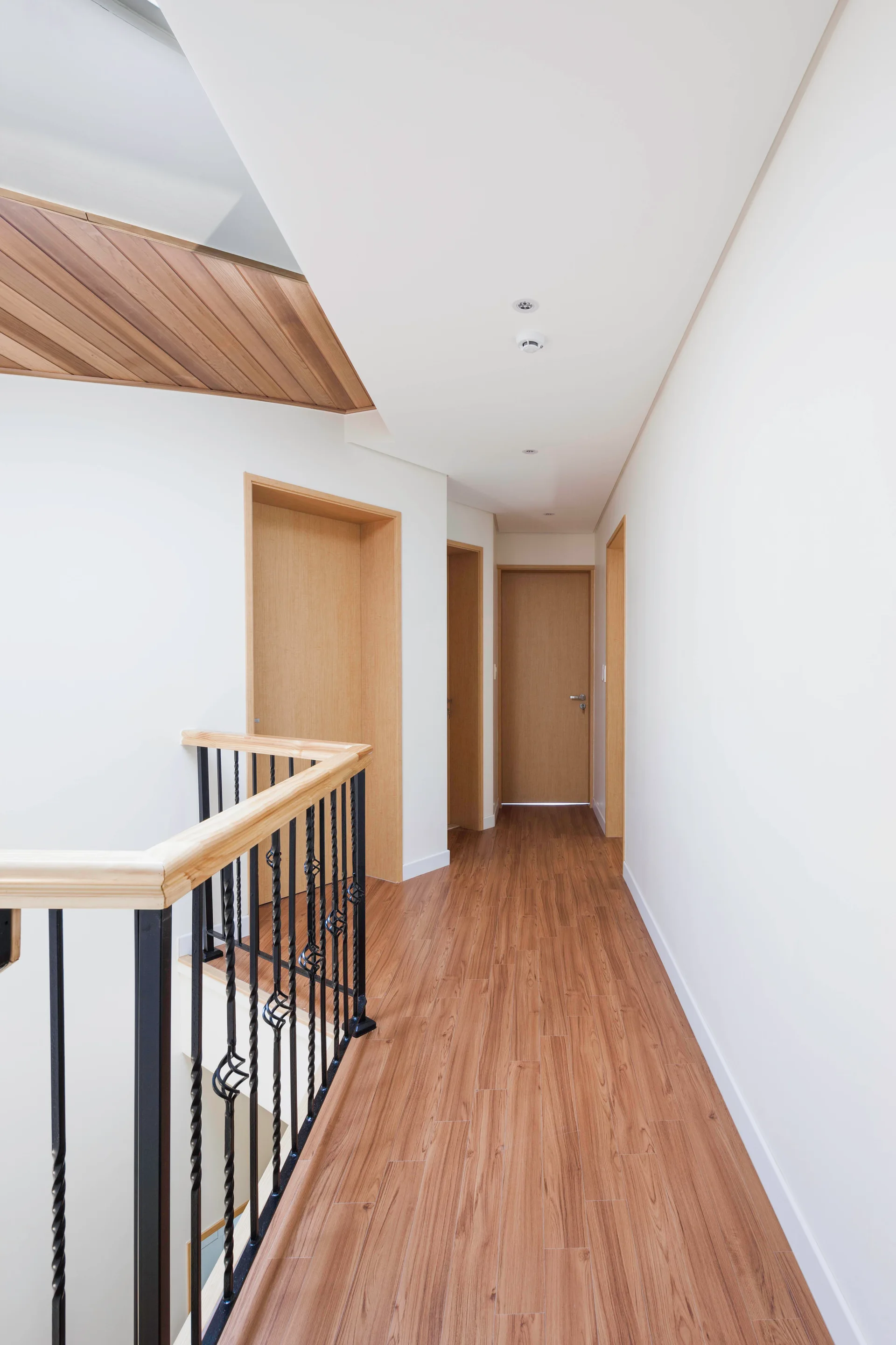 A modern second-floor hallway features warm wood flooring, matching flush doors, crisp white walls, and a black staircase railing.