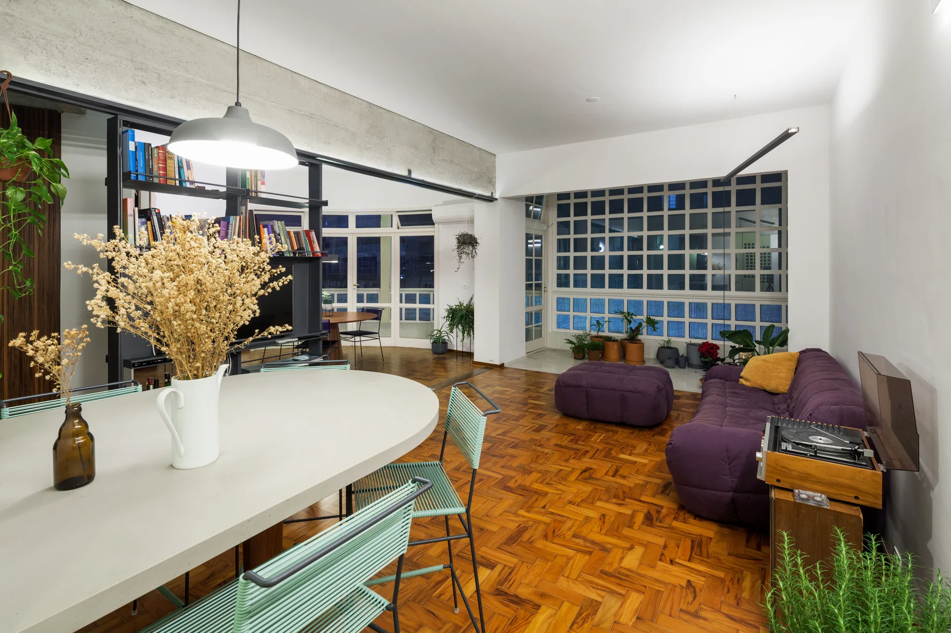 An open-concept room features a light oval dining table, a low-profile purple sofa, herringbone wood flooring, and an exposed concrete beam.