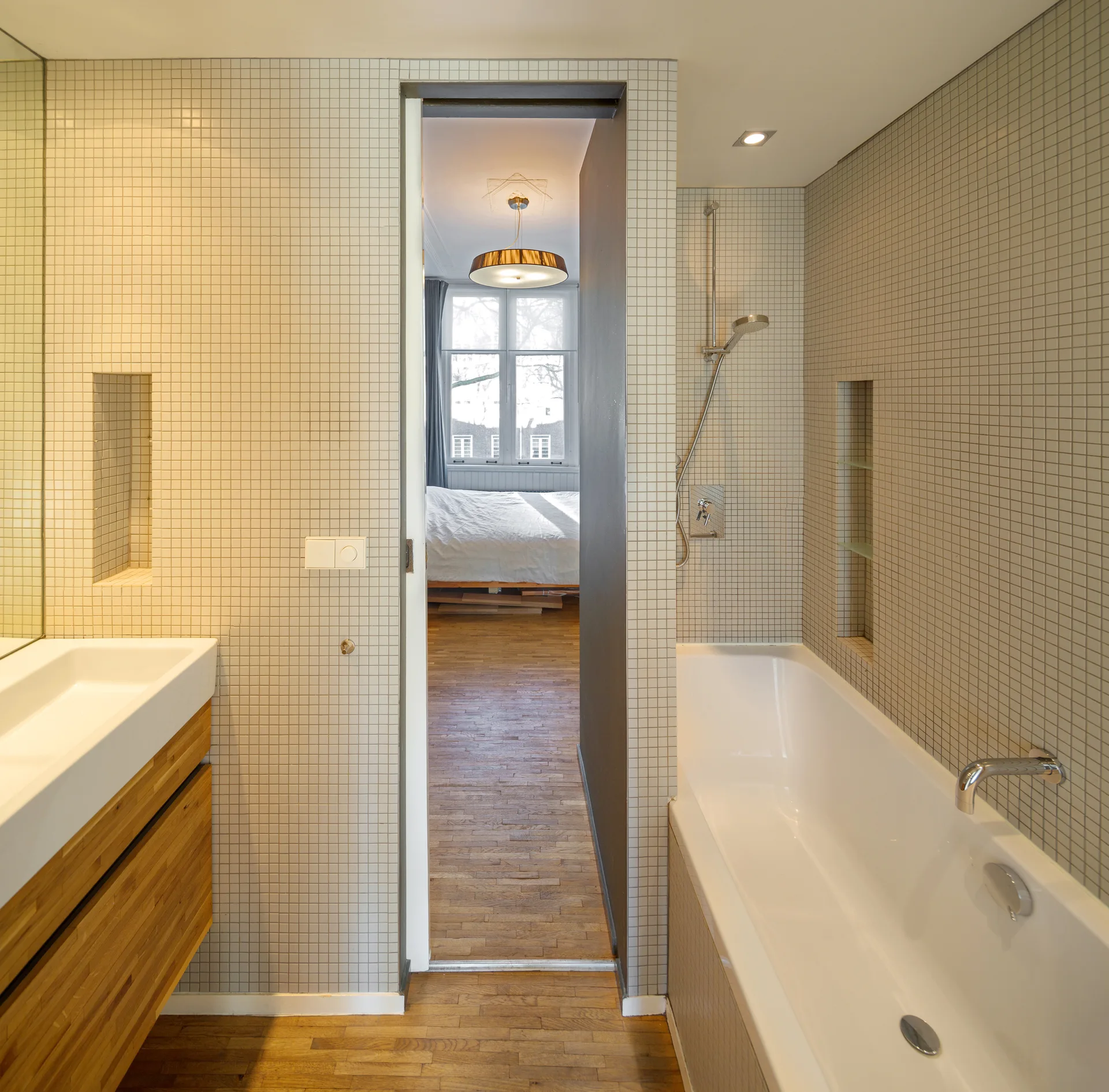 A modern bathroom with beige grid mosaic tiled walls, a white bathtub, a floating wood-toned vanity, and an open doorway looking into a bedroom.