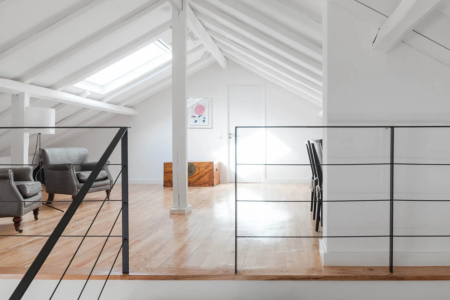 An open attic landing with light wood floors, a white sloped ceiling with exposed beams, and a modern black metal railing.