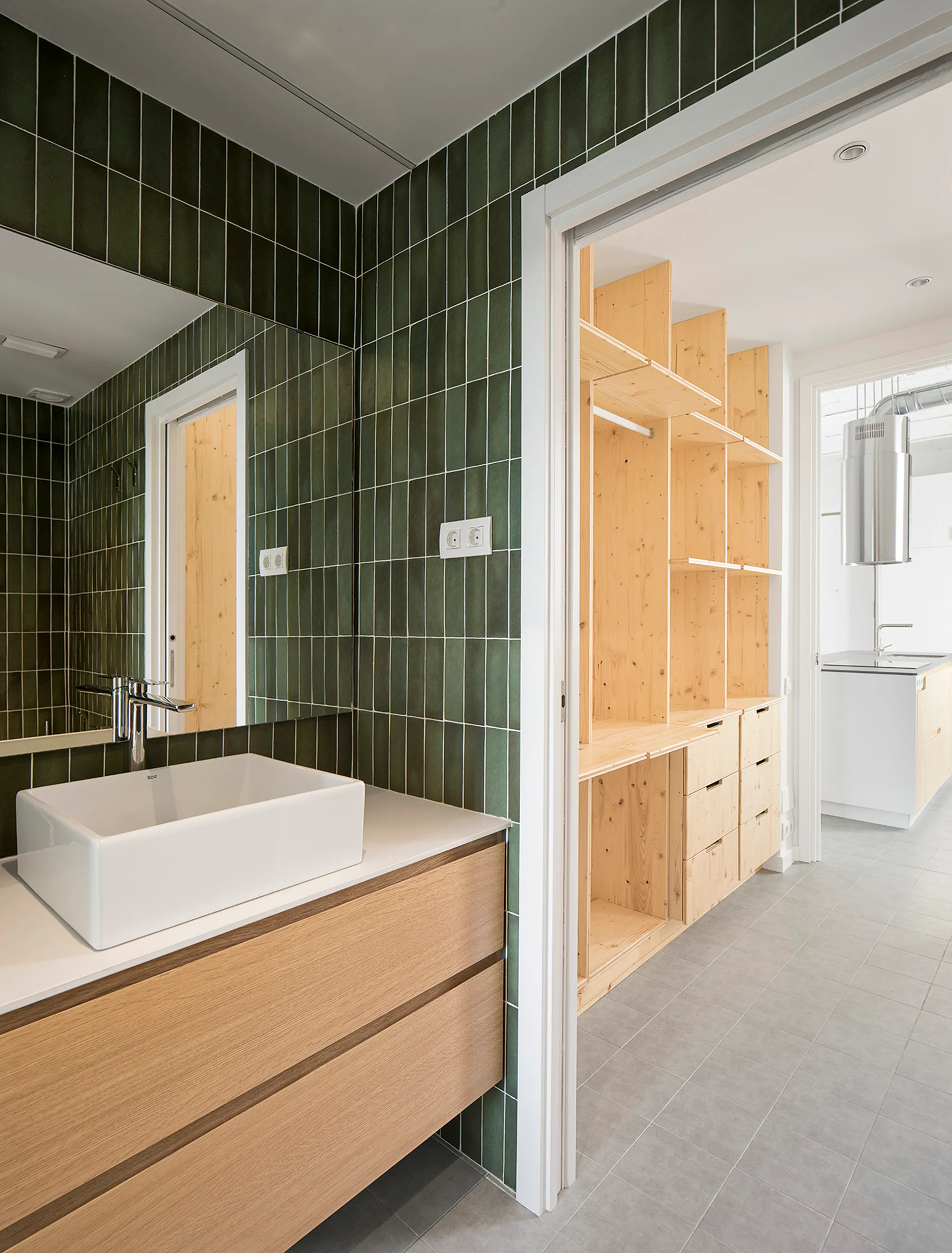 A modern bathroom features forest green vertically stacked wall tiles, a light wood vanity with a white vessel sink, and adjacent natural pine shelving.