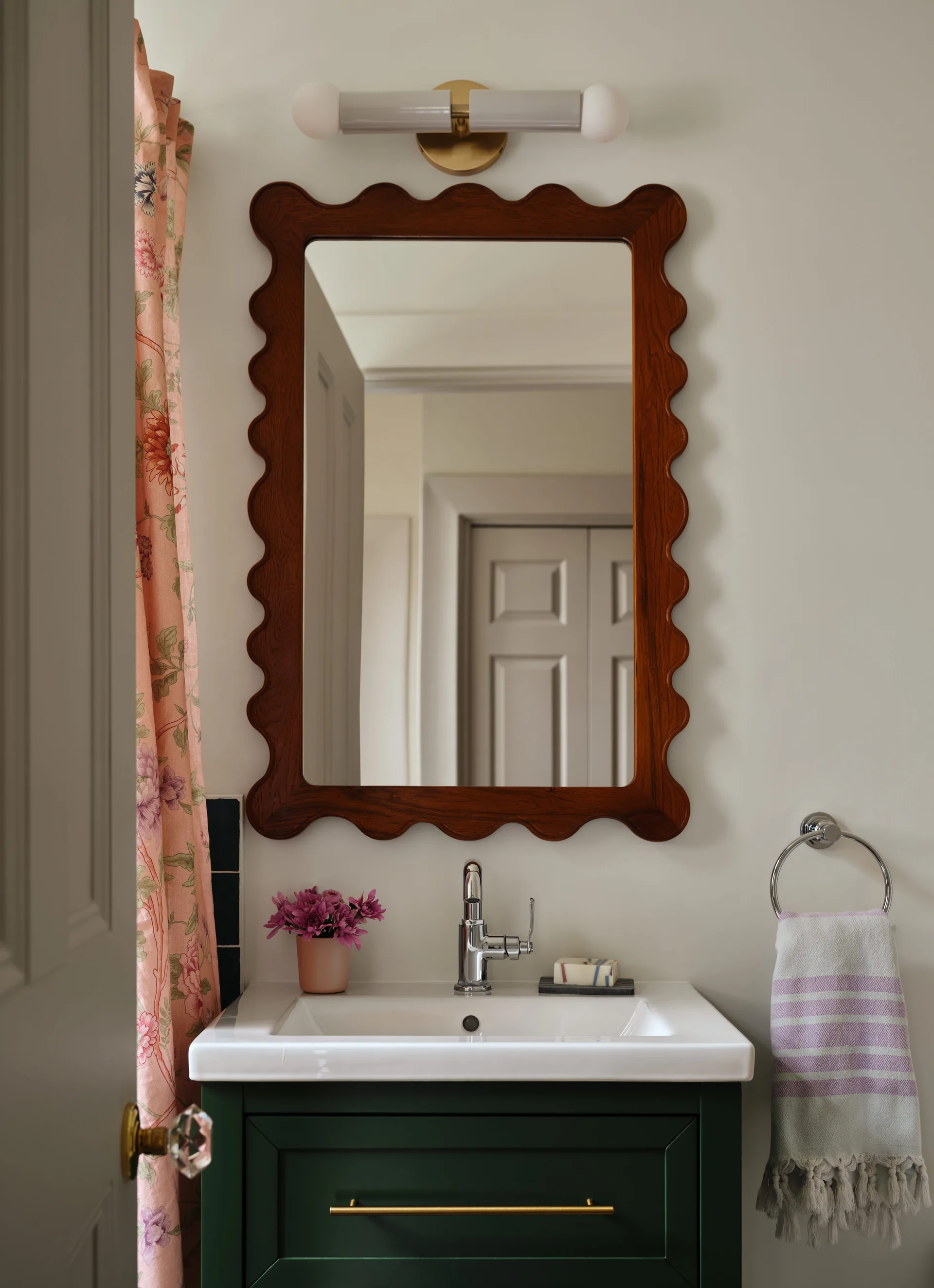 A bathroom vanity featuring a dark green cabinet, white ceramic sink, scalloped wood-framed mirror, and floral patterned curtain.