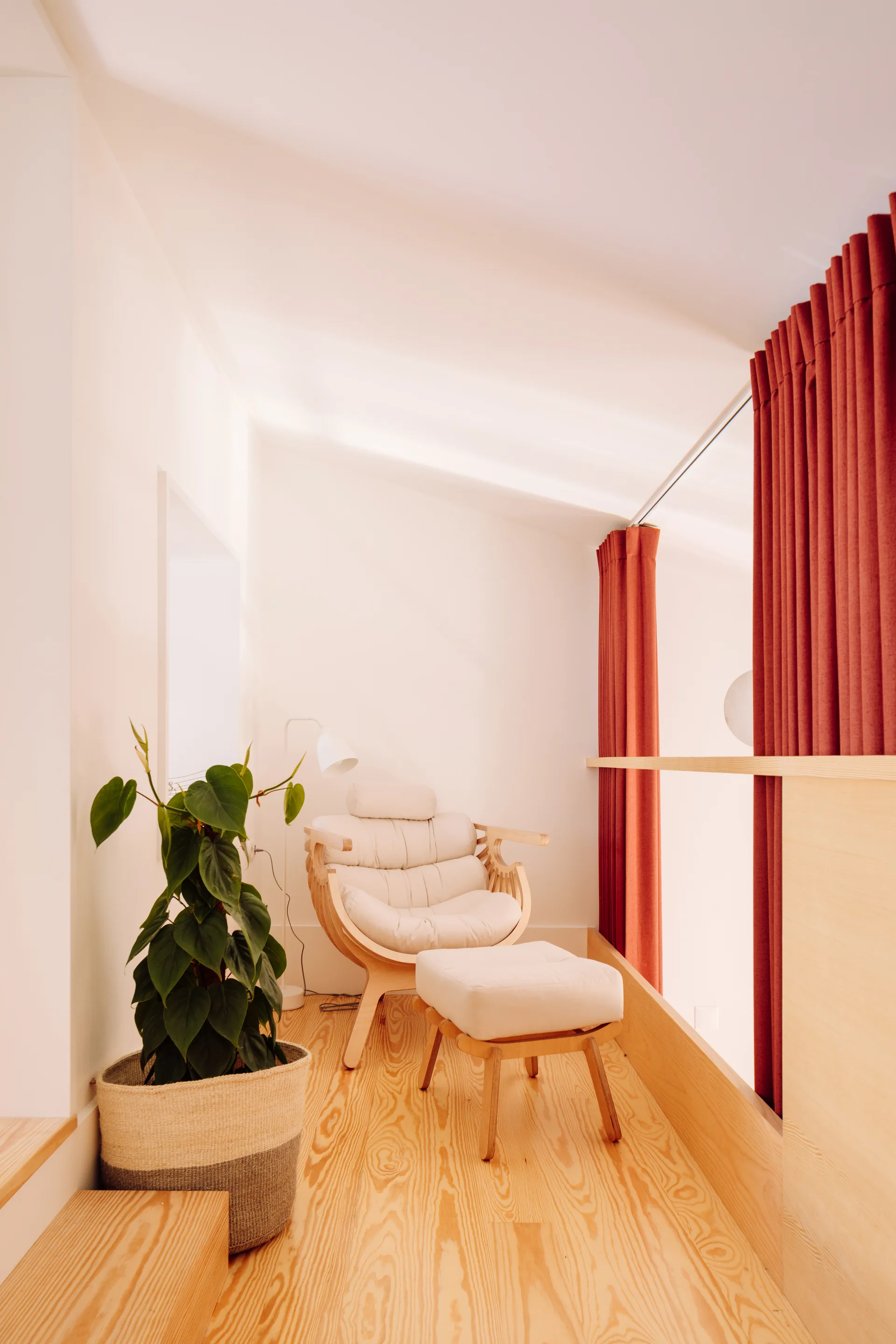 A cream upholstered lounge chair and ottoman sit on a light wood floor next to a green potted plant and red floor-length curtains on a loft landing.