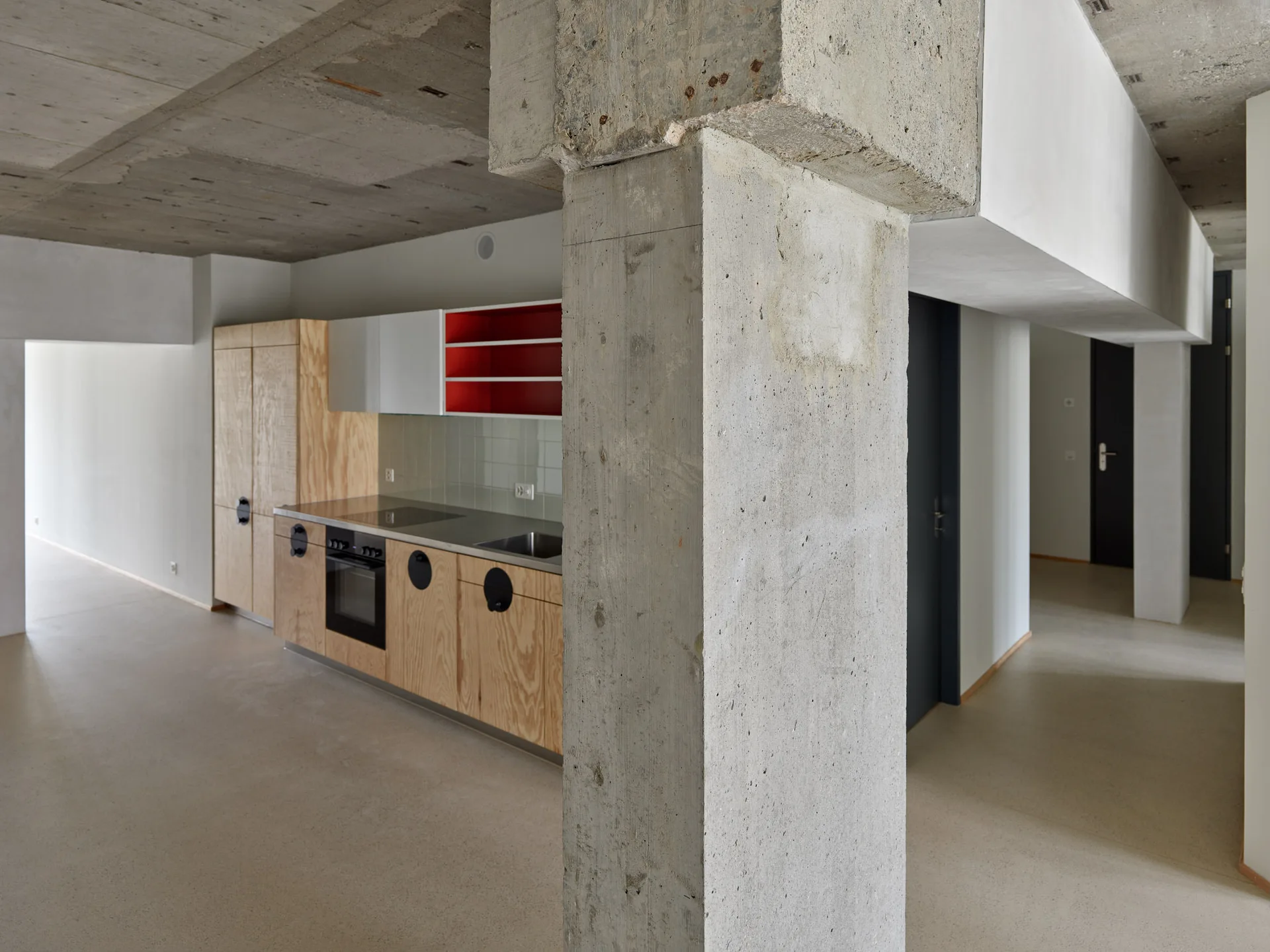 A minimalist kitchen features plywood base cabinets with black circular handles, a stainless steel countertop, and a prominent raw concrete column.