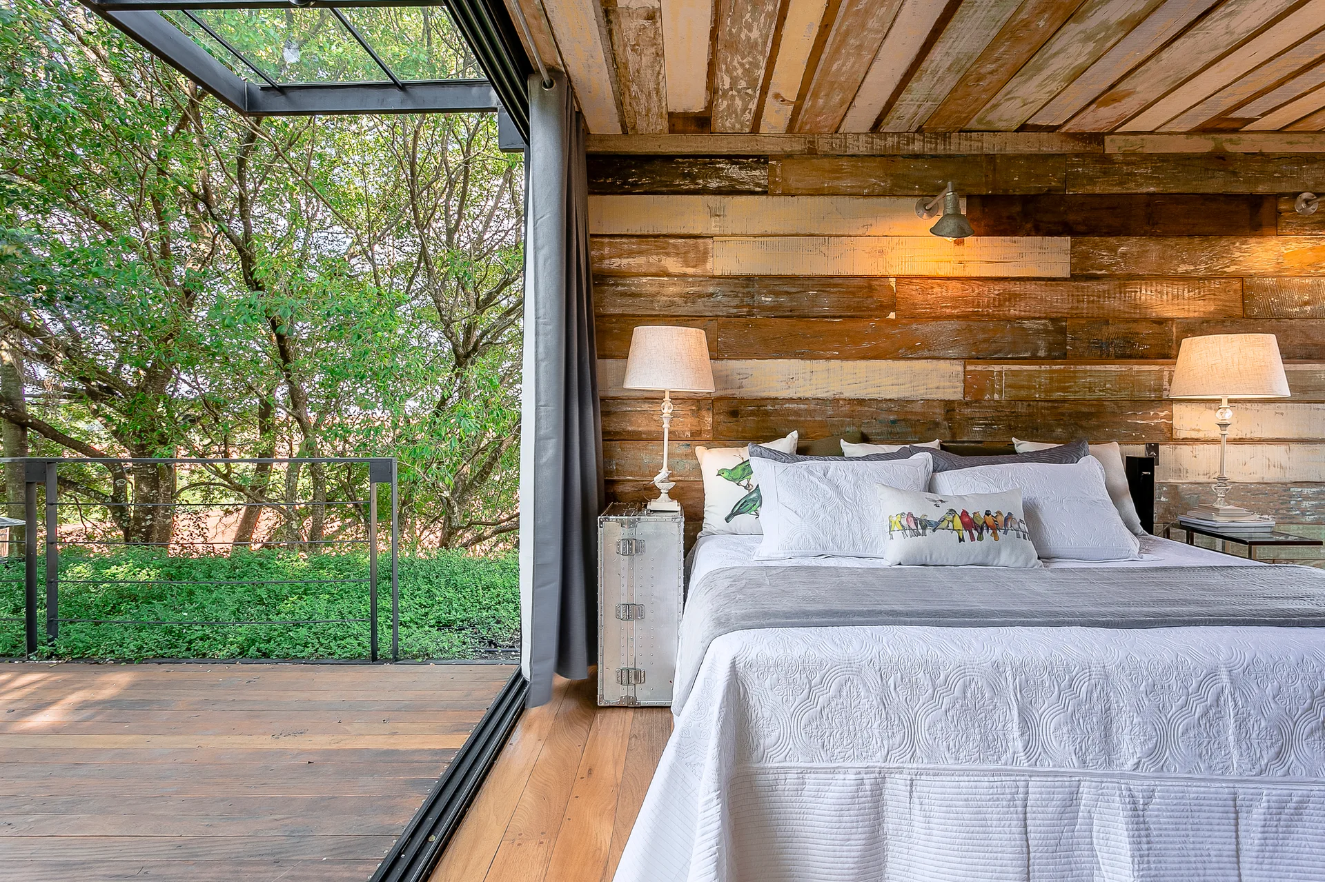 A rustic bedroom features a bed with white linens against a reclaimed wood wall next to an open sliding door leading to a balcony.