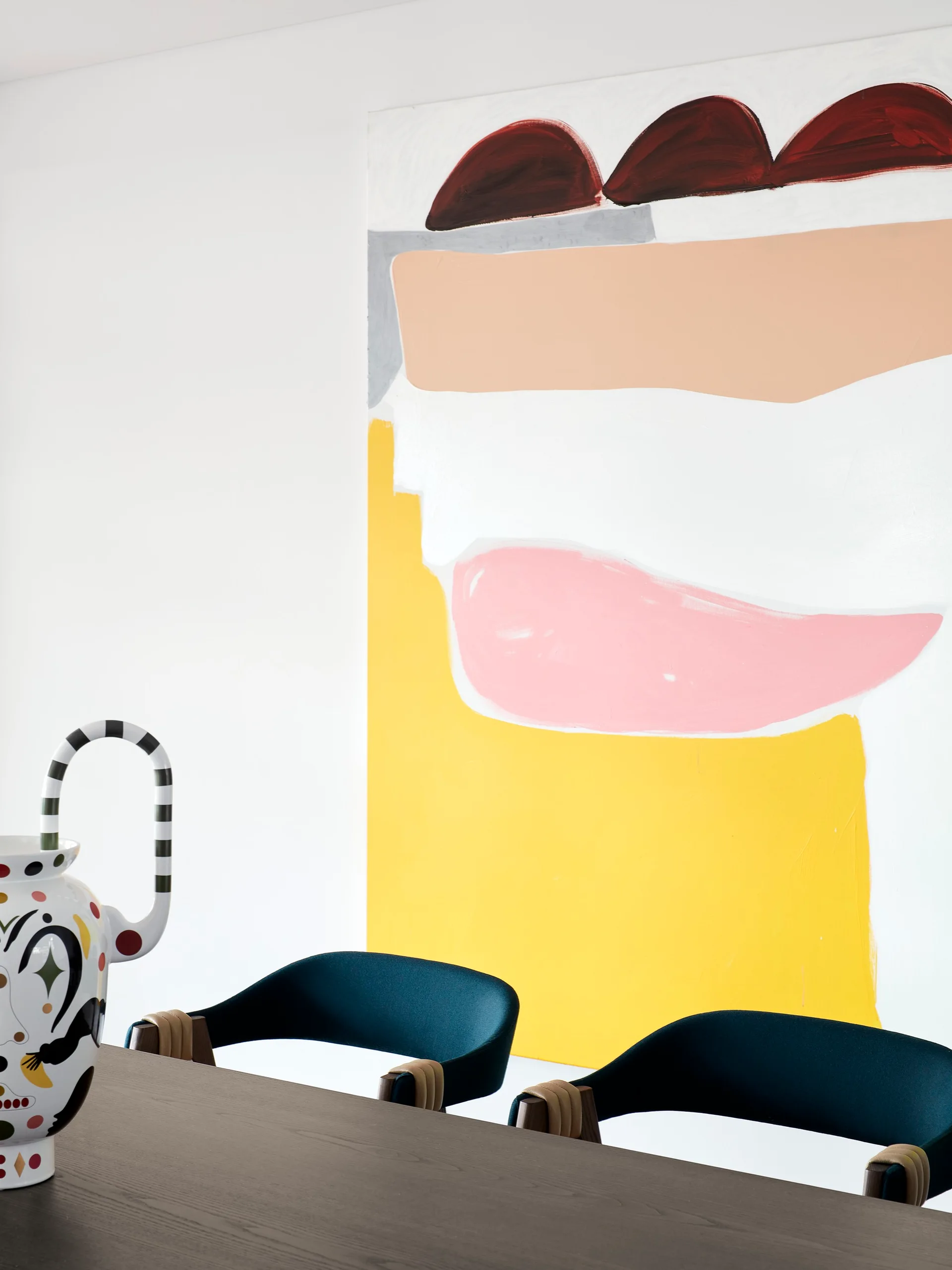 Two dark upholstered dining chairs sit at a dark wooden table in front of a white wall decorated with a large, colorful abstract painting.