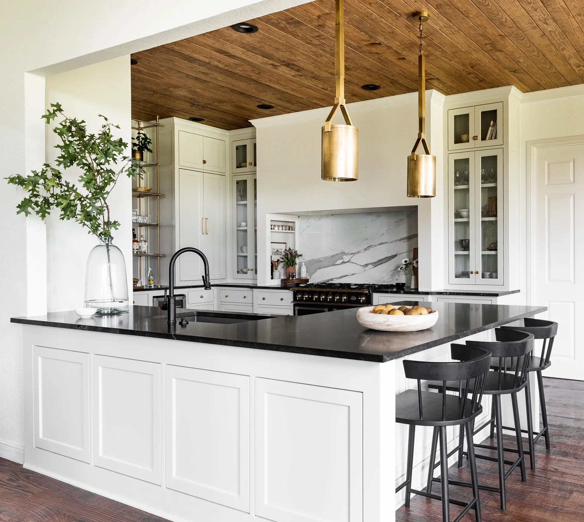 A kitchen featuring white cabinetry, a black-topped island with matching black stools, brass pendant lights, and a warm wood plank ceiling.