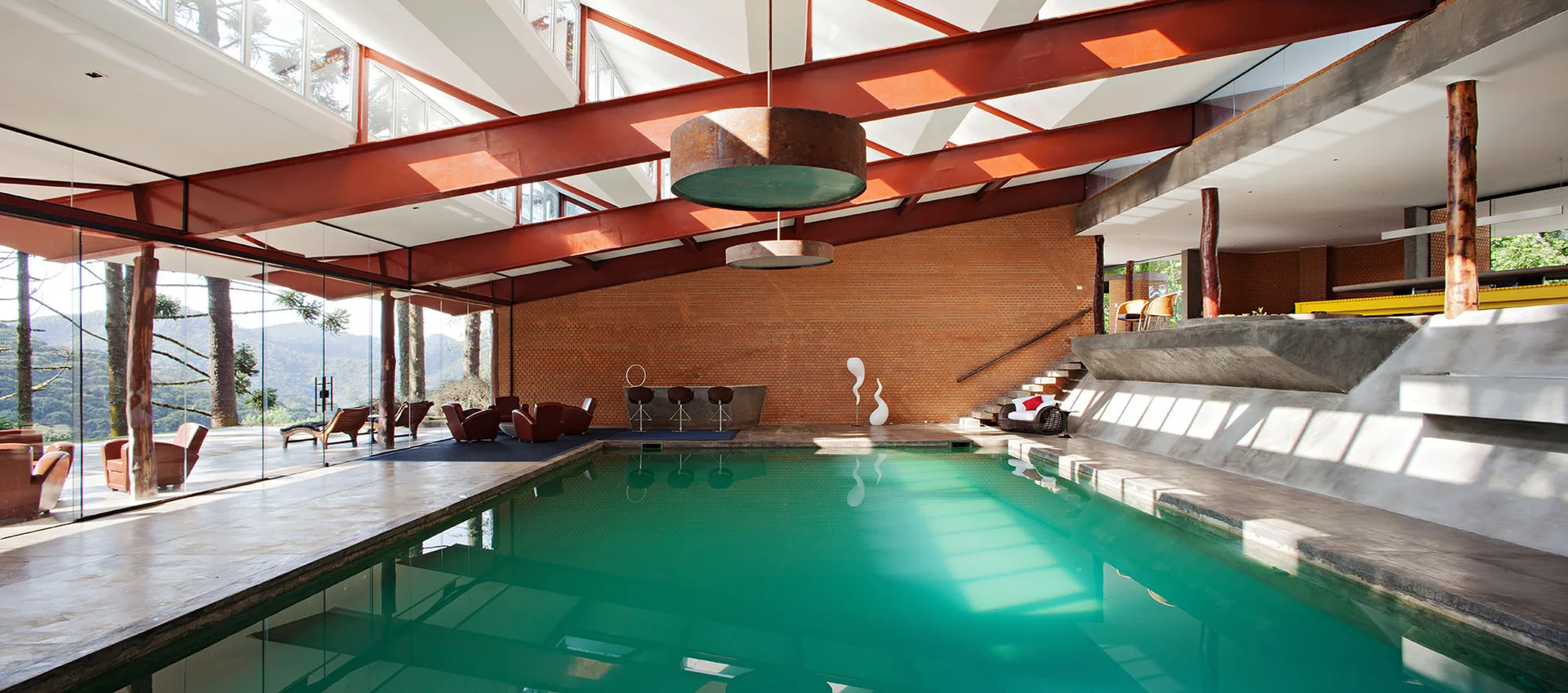 A large indoor swimming pool with turquoise water, a red-painted structural steel ceiling, and an exposed brick wall.