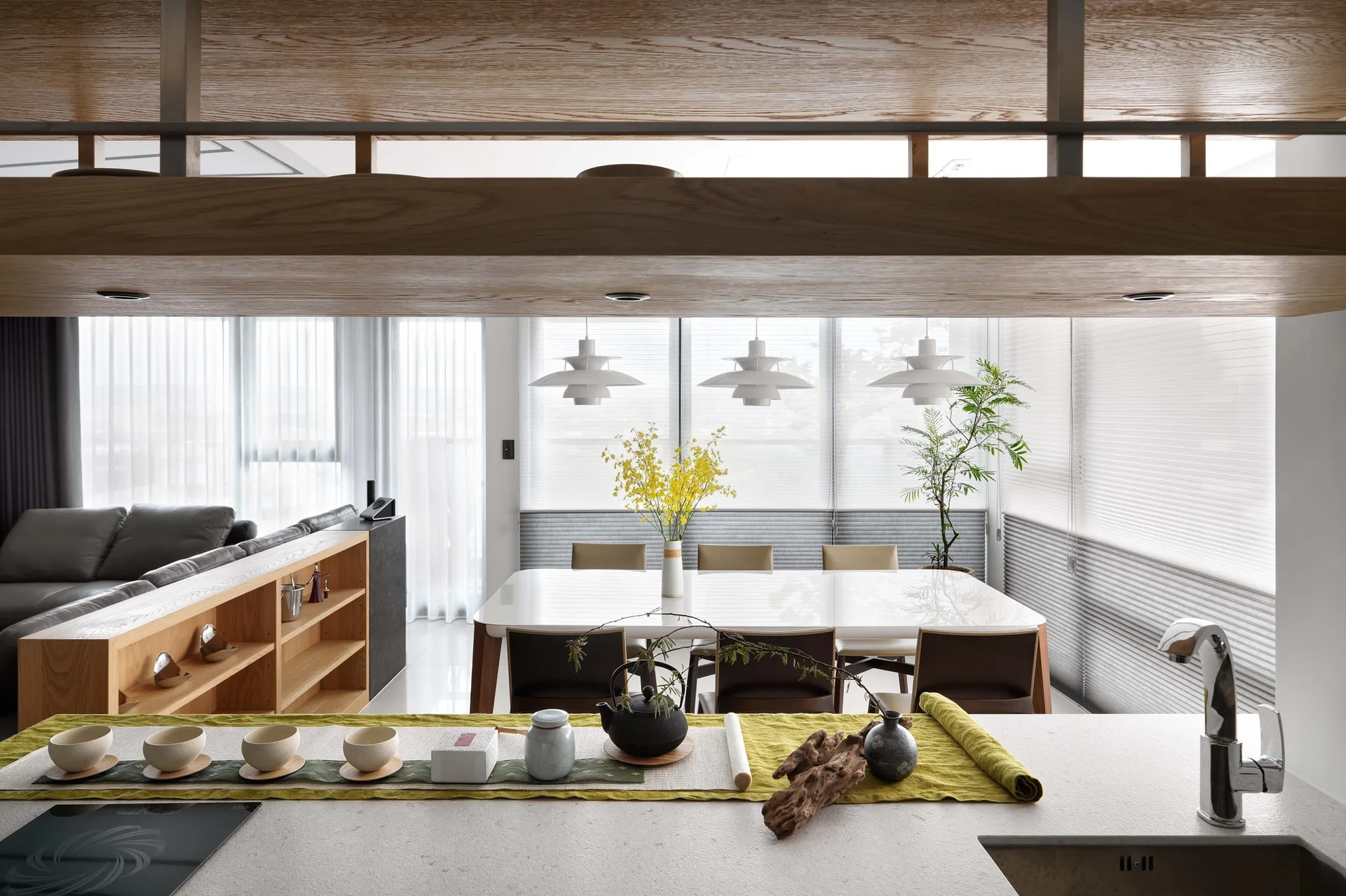 A modern kitchen counter with a tea set looks out toward a dining table with three white pendant lights and a grey sofa in the background.