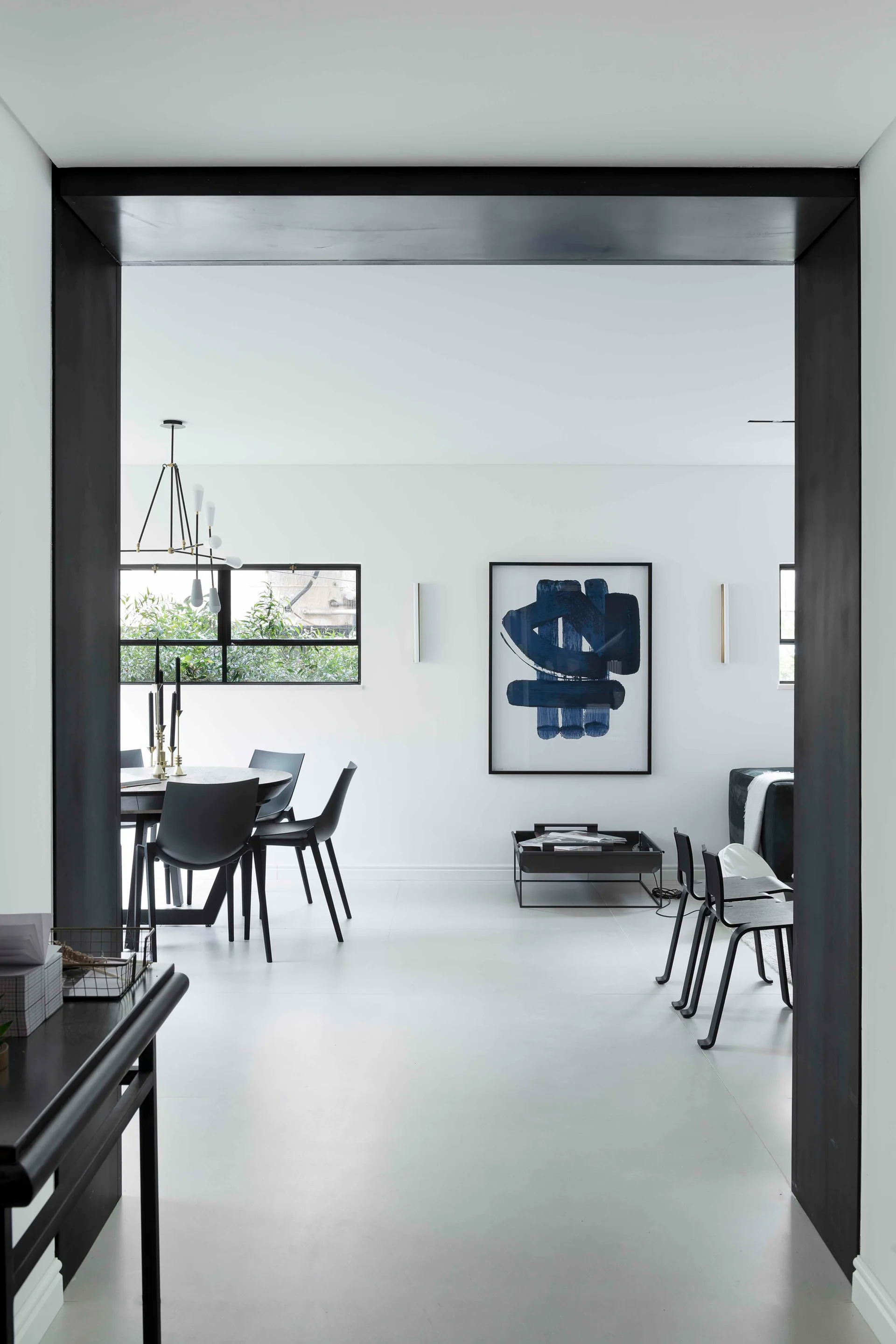 A minimalist dining room with black chairs and a round table seen through a black doorway, featuring a large blue abstract painting on a white wall.