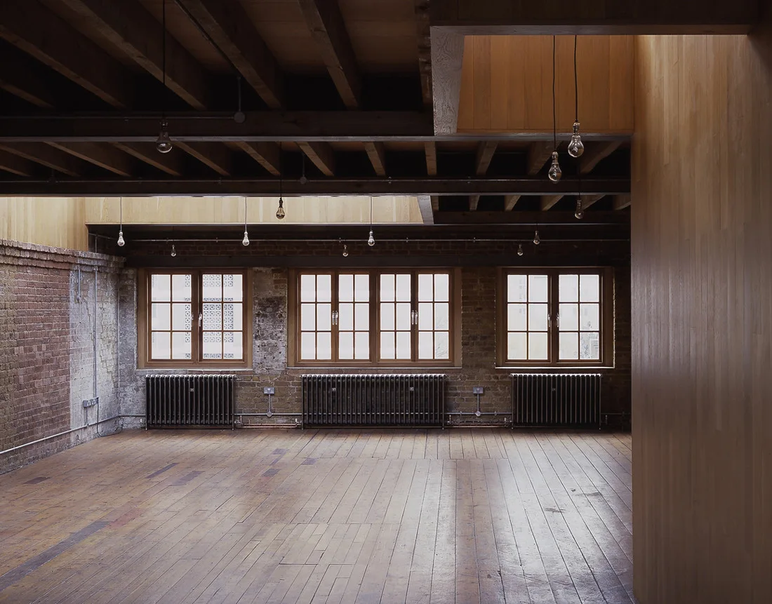 An empty industrial loft features wide wood floorboards, an exposed brick wall, three windows with black column radiators, and bare bulb pendant lights.