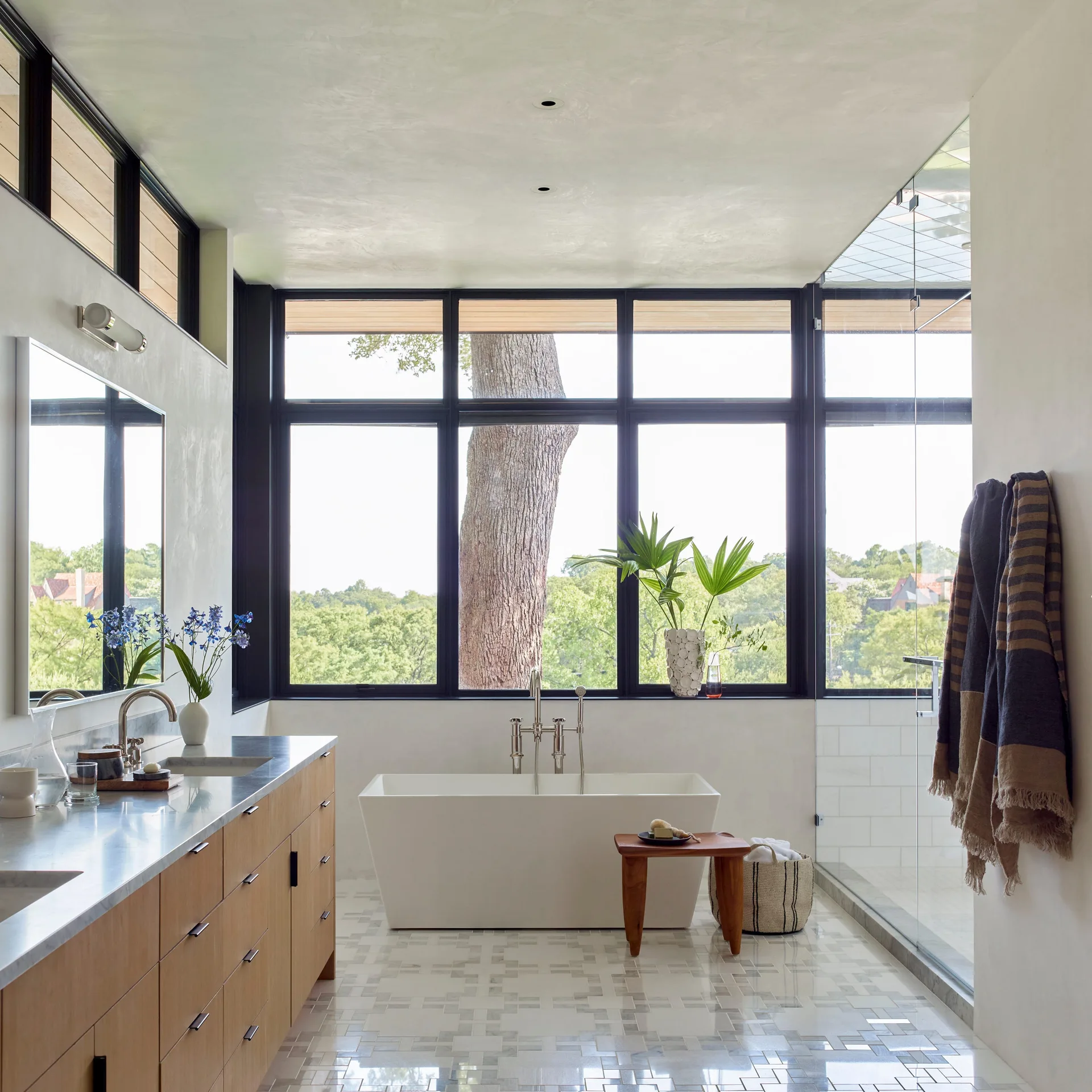 A bright modern bathroom features a freestanding white bathtub in front of a large window, a wood vanity, and a geometric tiled floor.