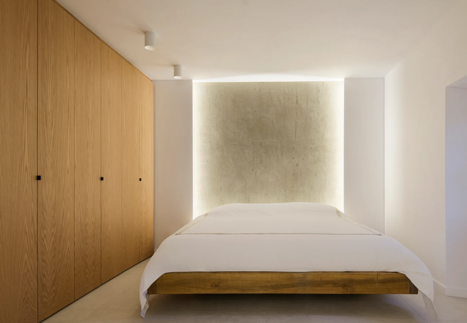 A low wooden platform bed with white bedding sits before a textured plaster accent wall illuminated by indirect cove lighting next to an oak wardrobe.
