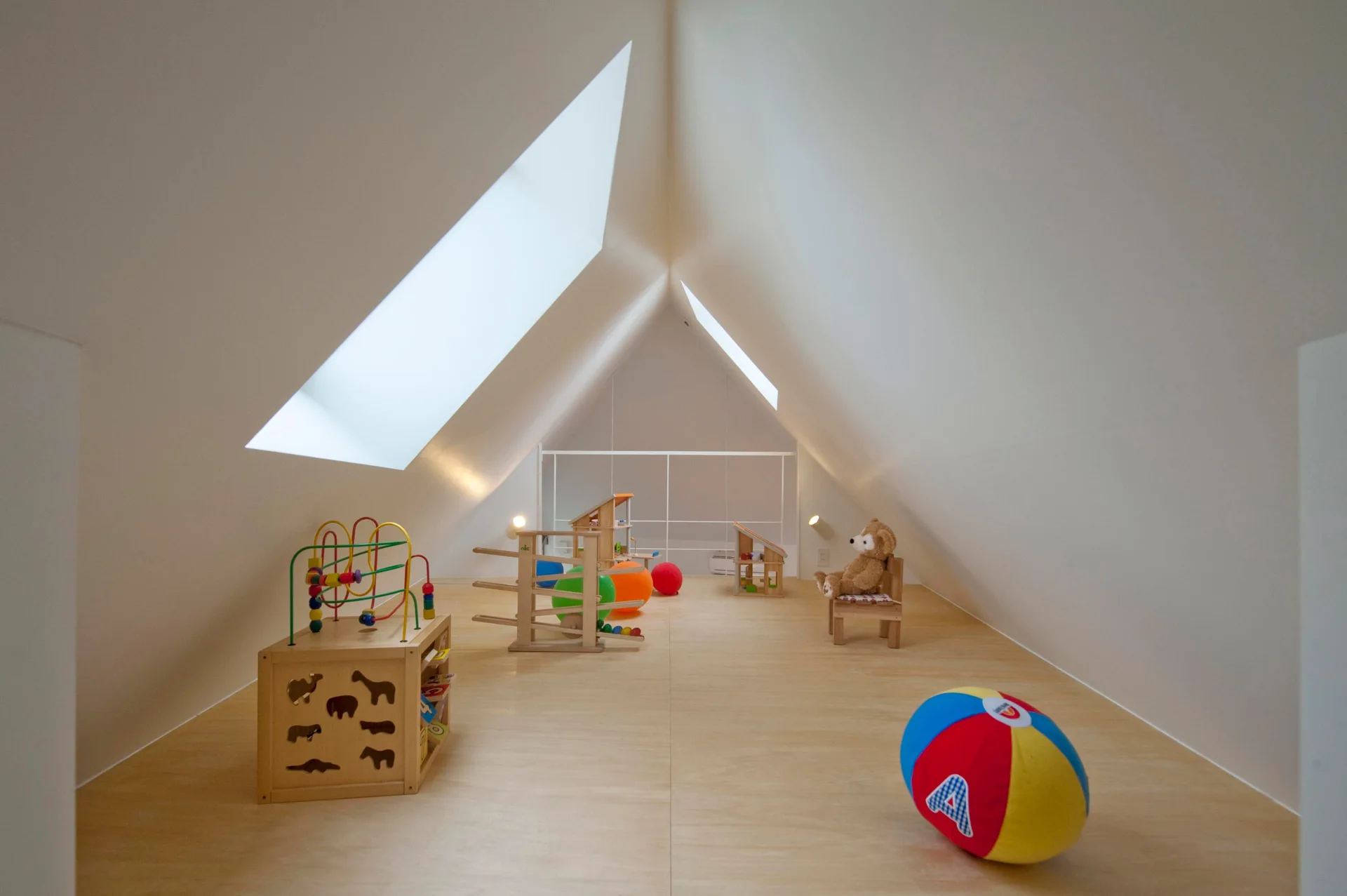 A minimalist attic playroom features clean white sloped walls, dual skylights, a light wood floor, and simple wooden toys.