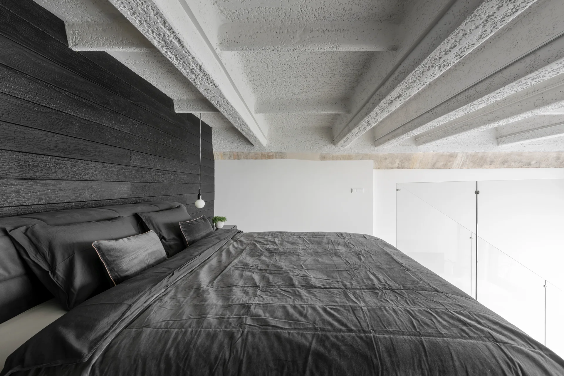 A minimalist loft bedroom showing a dark grey bed against a black wood paneled wall beneath textured white concrete ceiling beams.