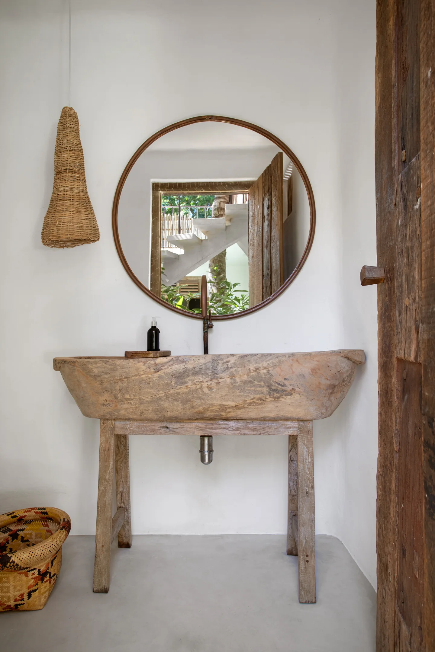 A rustic bathroom features a carved wooden trough sink, a large round mirror, a woven pendant light, and a concrete floor.
