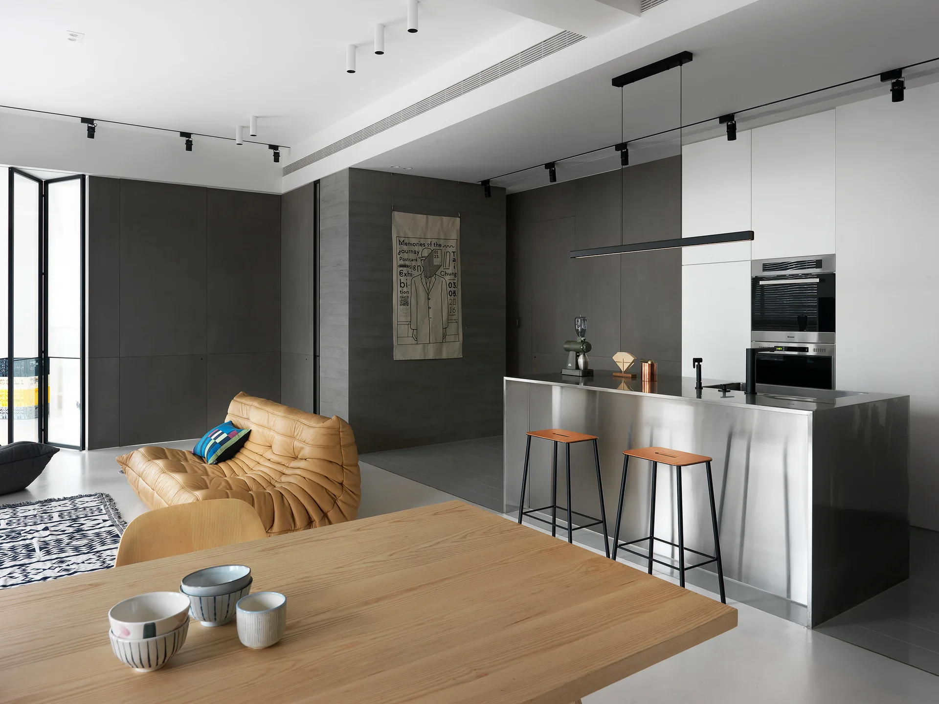 A modern open-plan kitchen and living space featuring a stainless steel island, a tan tufted leather sofa, and dark gray paneled walls.