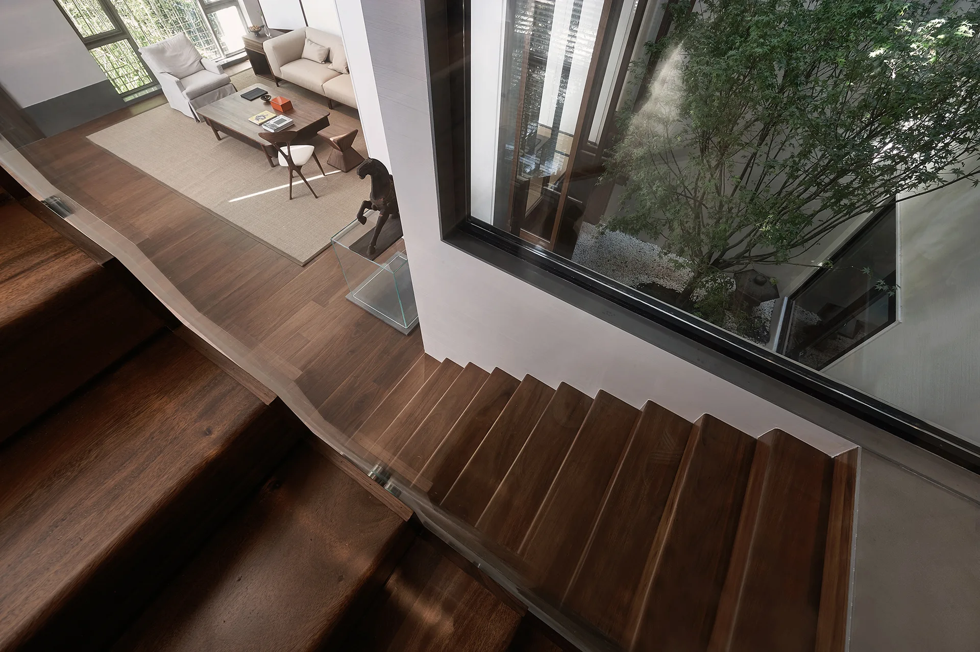 High-angle view of a wooden staircase with a clear glass railing next to a living room and an internal courtyard featuring a green tree.