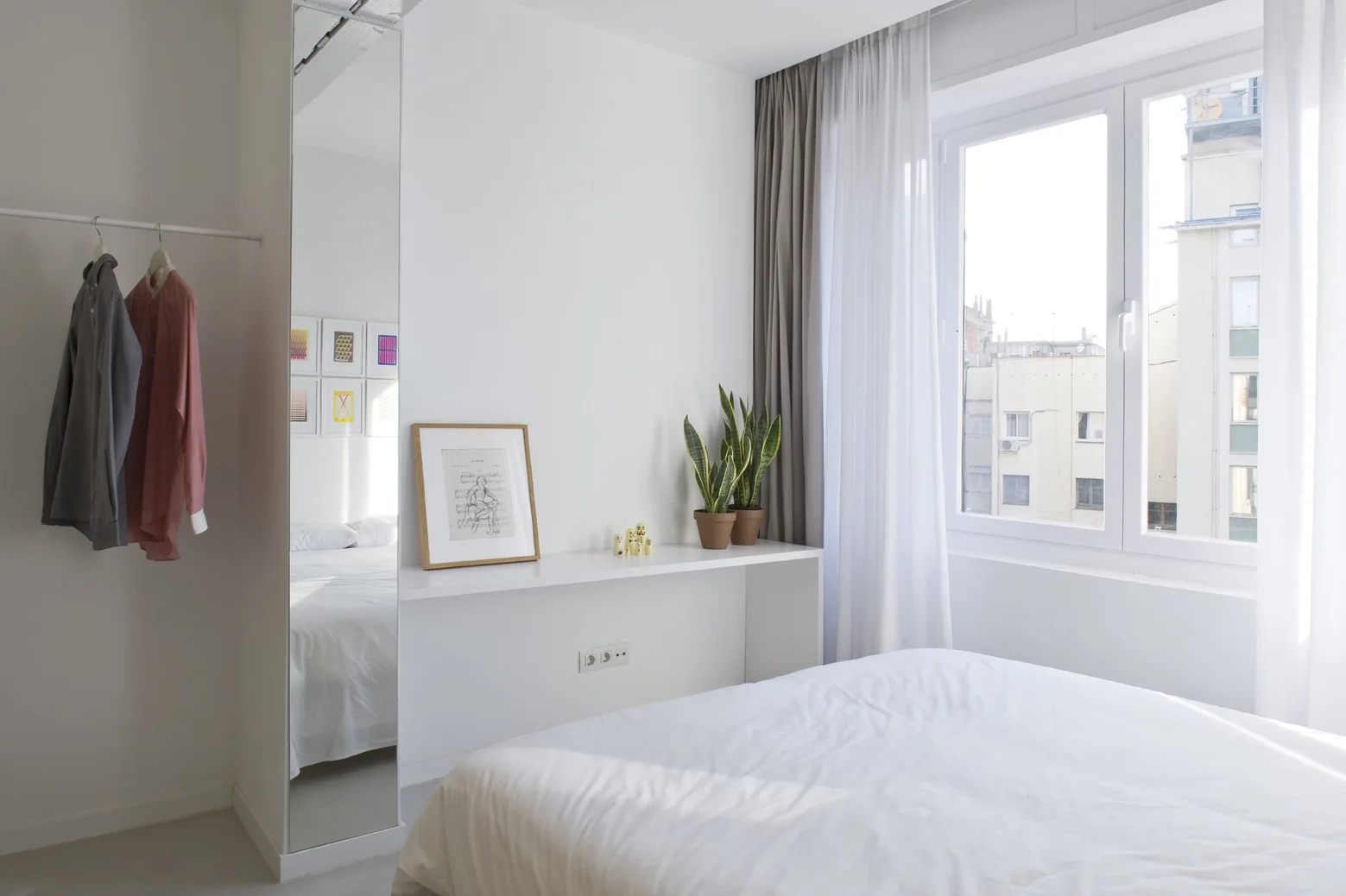 A minimalist bedroom with white walls, a bed with white bedding, a full-length mirror, a floating console shelf, and a clothing rail.