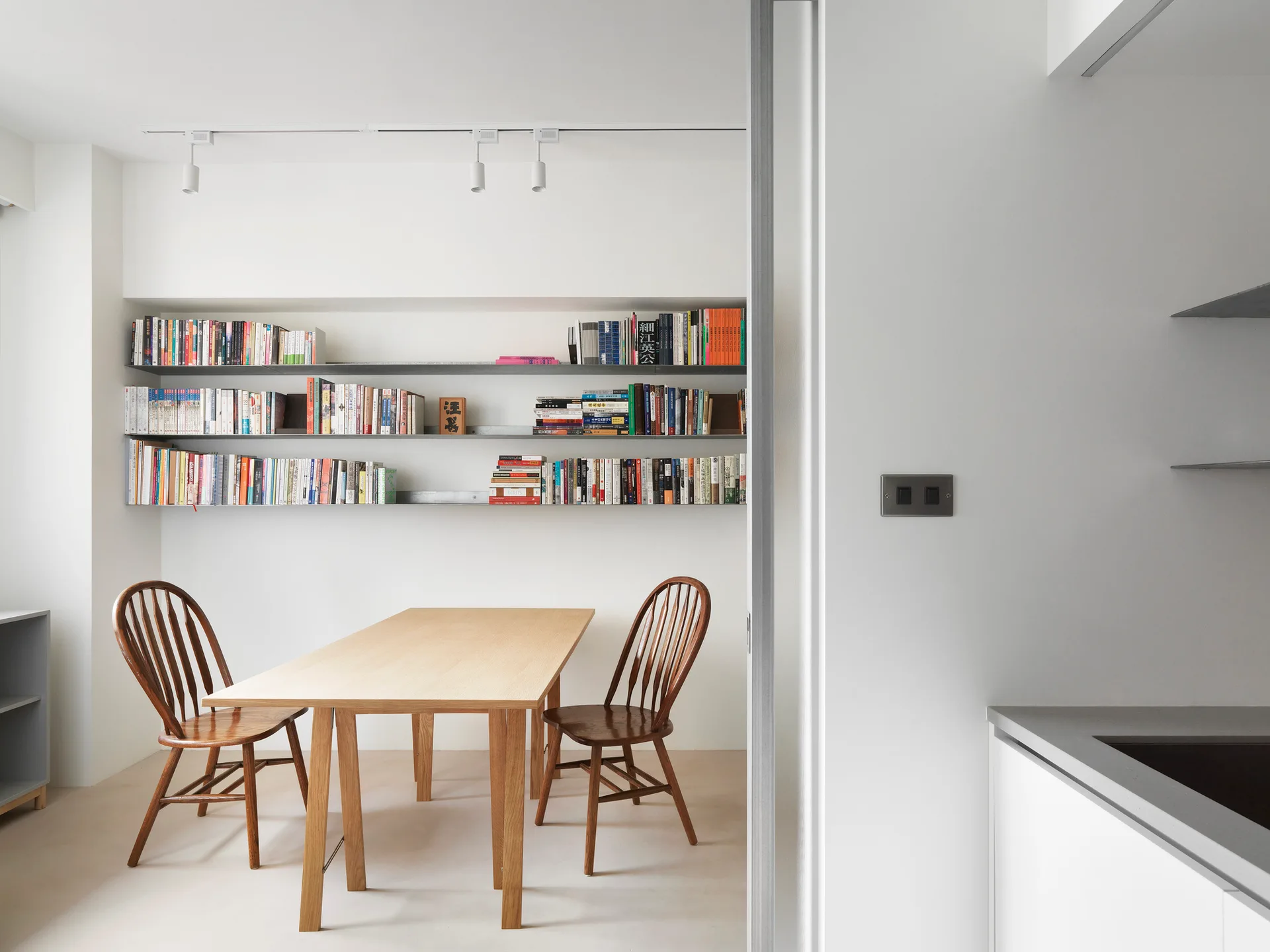 A minimalist dining room features a light wood table, two brown wooden chairs, and three long wall-mounted bookshelves filled with books.