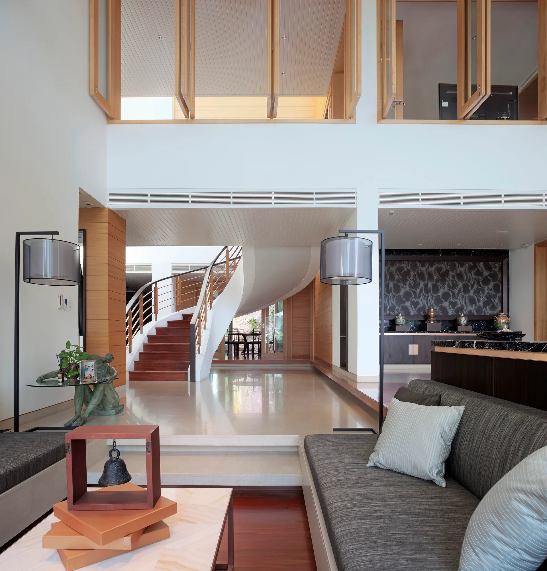 A contemporary sunken living room featuring gray sofas, thin floor lamps, and a polished floor leading to a curved white staircase with wood steps.