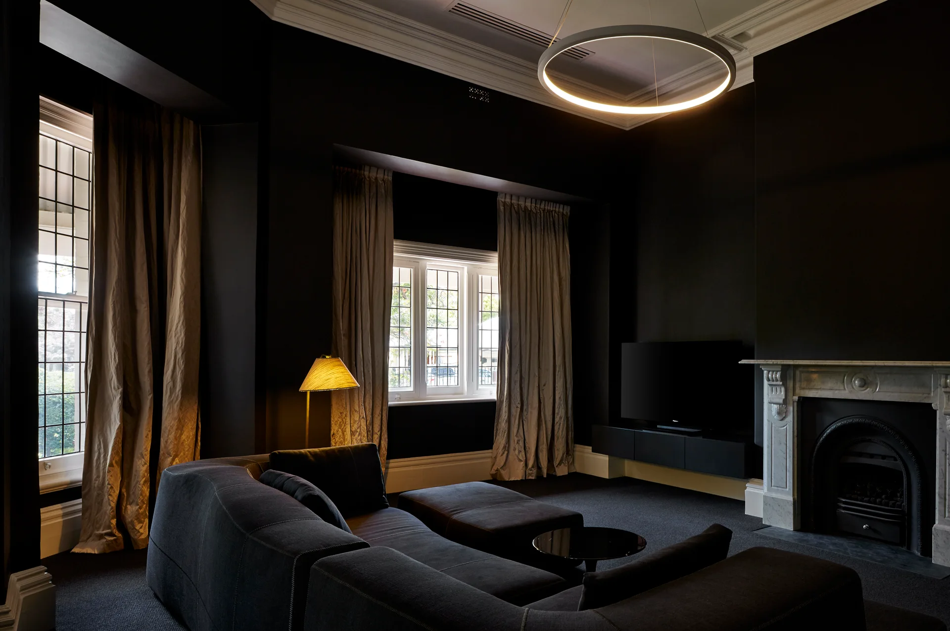 A moody living room features dark charcoal walls, a large black sectional sofa, a white marble fireplace, and a circular halo pendant light.