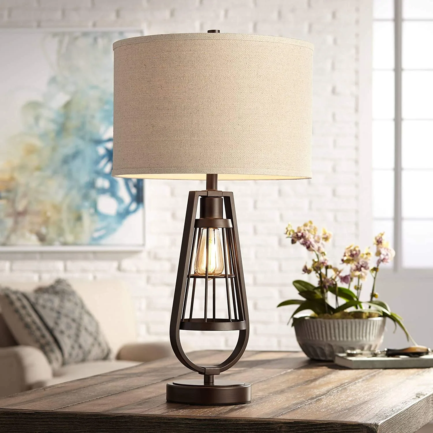 A bronze cage-style table lamp with a lit bulb and beige drum shade sits on a wooden table against a white brick wall.