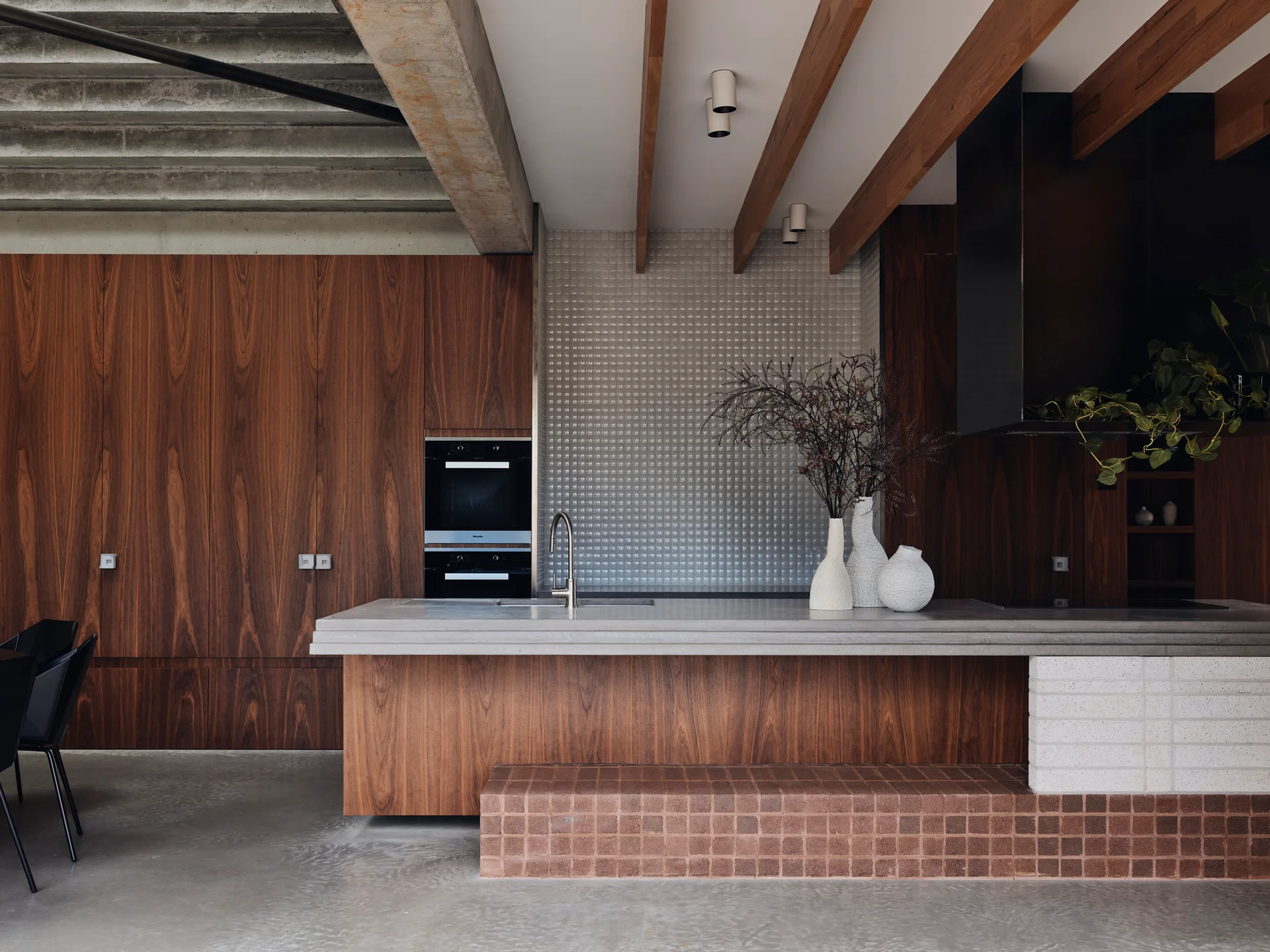 A modern kitchen features rich wood cabinetry, a concrete-topped island with a brown tile footrest, and a shimmering metallic grid backsplash.