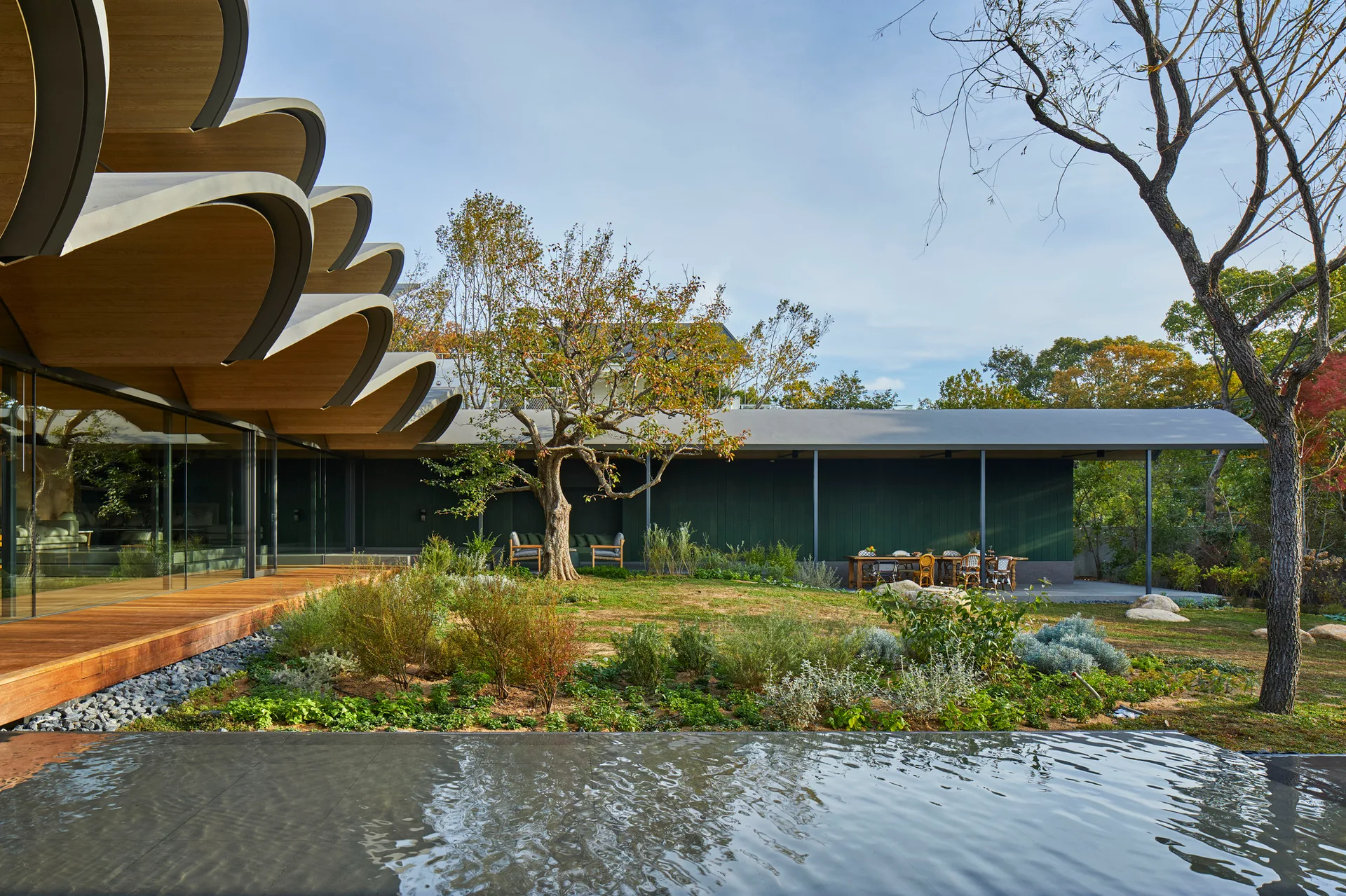 A modern house with a dramatic scalloped wood roof features a wood deck, a dark green exterior wall, and a central garden next to a reflective pool.