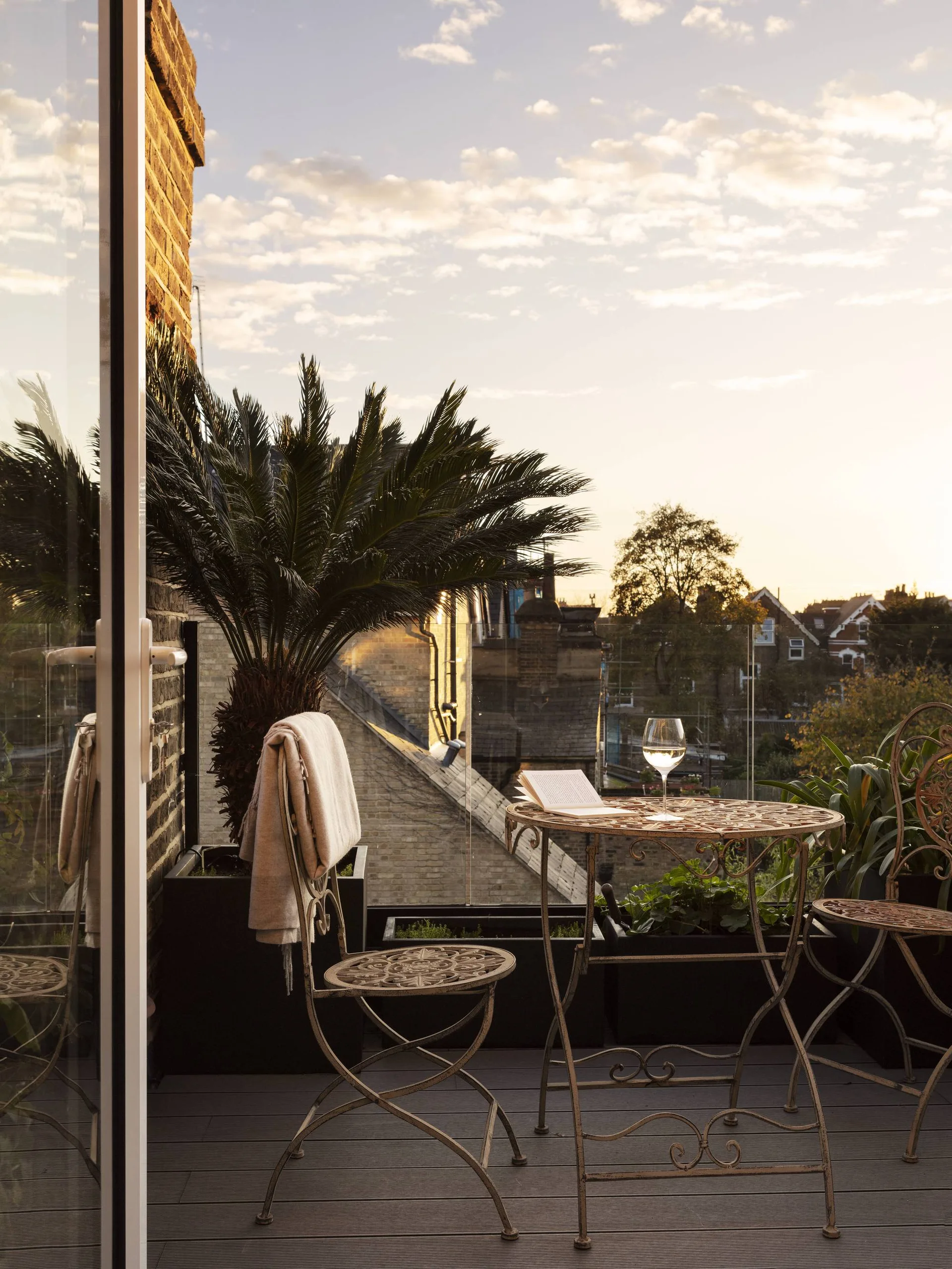 An ornate metal bistro set sits on a grey-planked balcony with a transparent glass railing, overlooking rooftops under a soft sunset sky.
