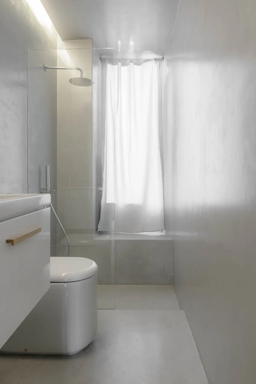 A narrow monochromatic bathroom features seamless light gray plaster walls, a white toilet, and a glass-enclosed shower with a sheer curtain.