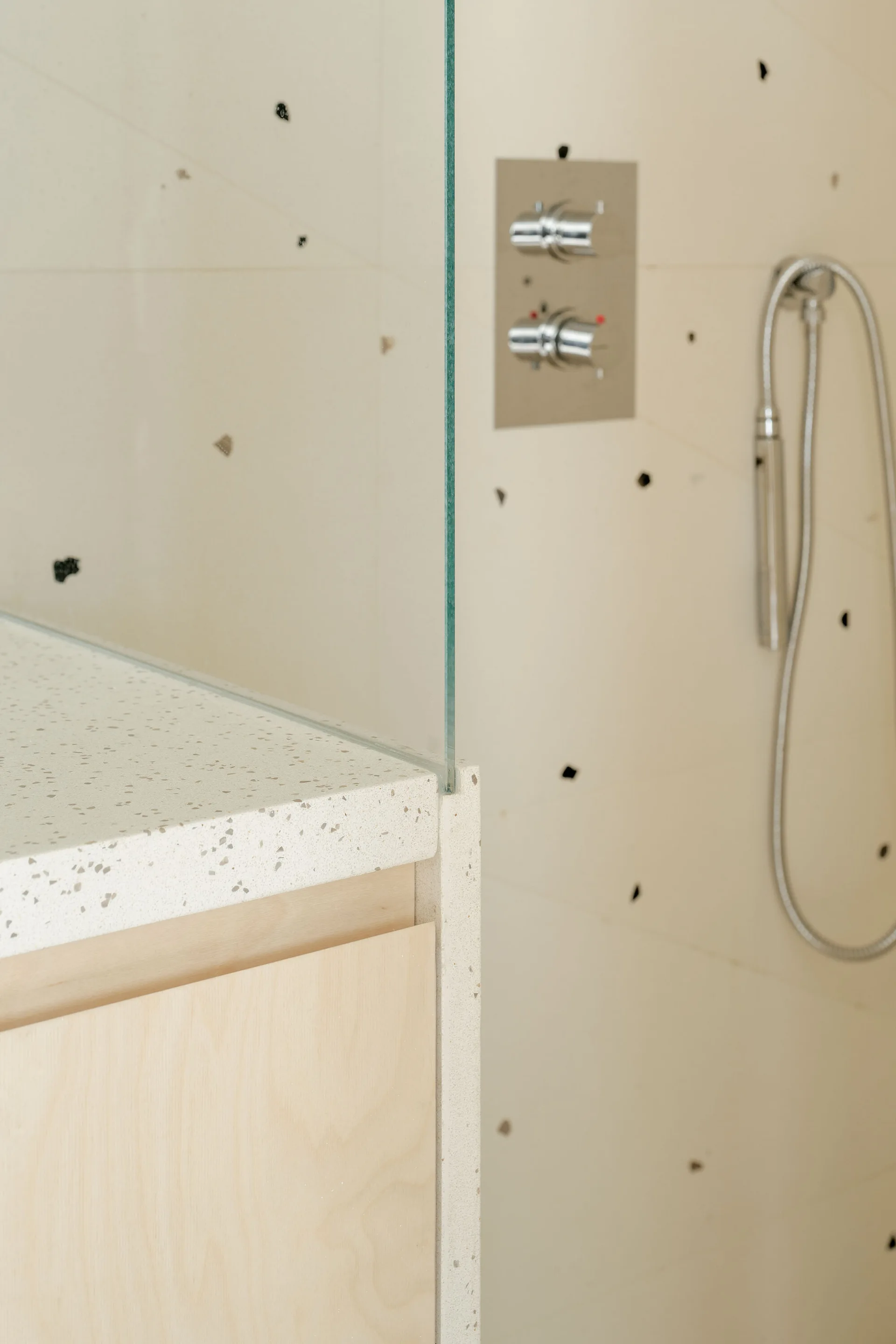 Close-up of a bathroom vanity with a speckled cream stone countertop, a glass shower partition, light wood cabinets, and chrome fixtures.
