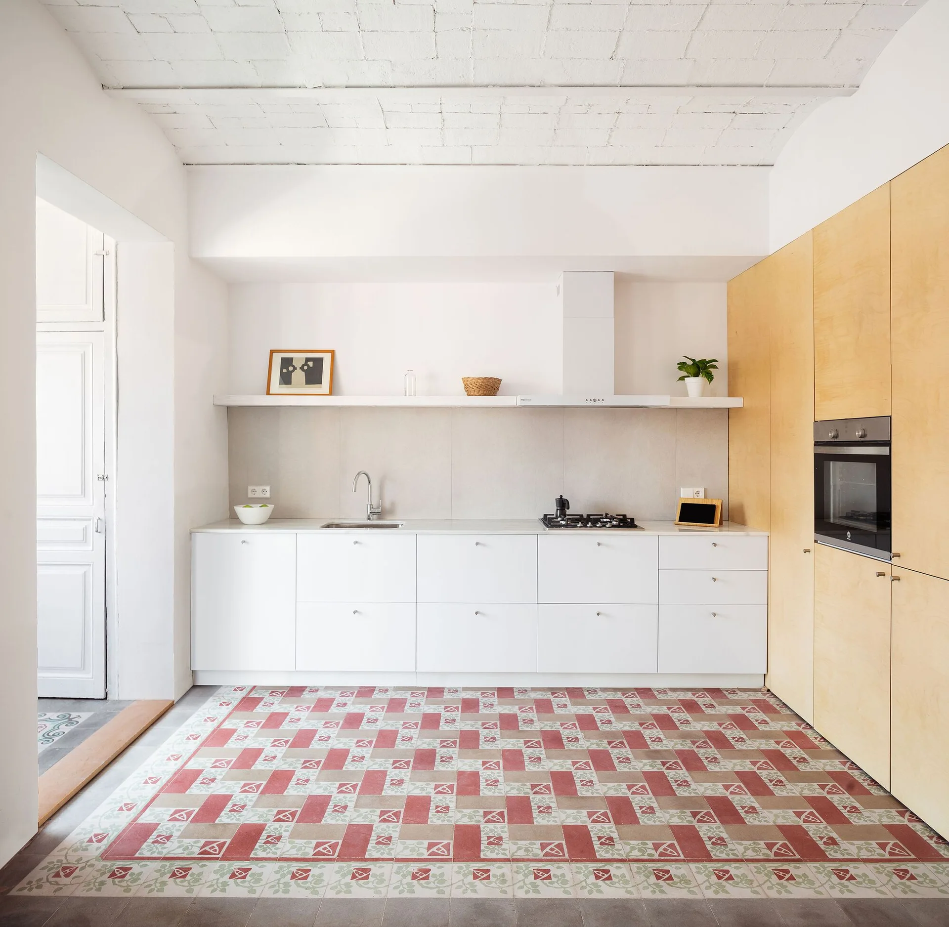 A modern kitchen featuring white lower cabinets, a full-height light wood pantry wall, a floating shelf, and red patterned floor tiles.
