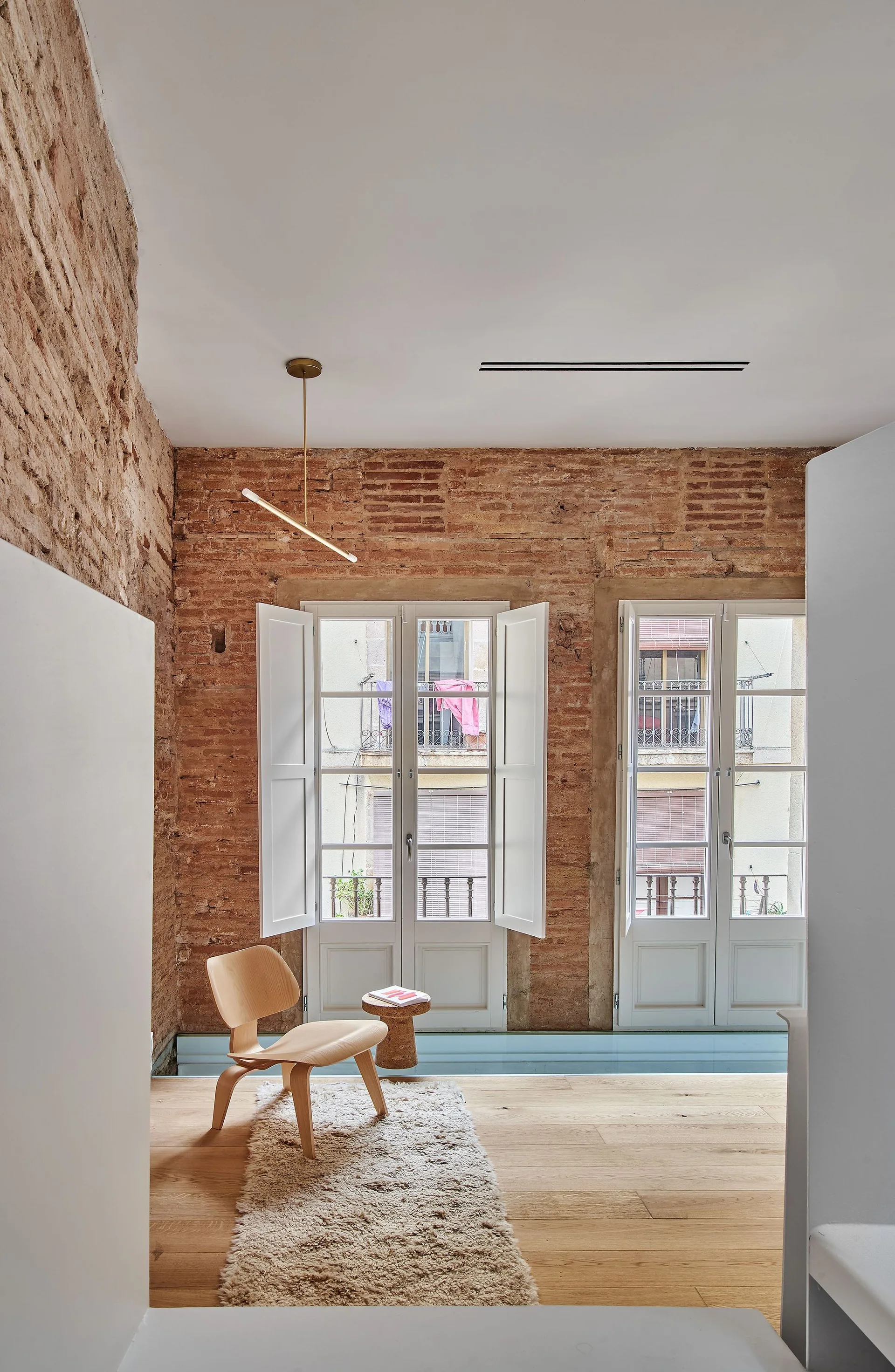 A minimalist room features an exposed brick wall, two tall windows with white interior shutters, a single wooden chair, and a small cork side table.