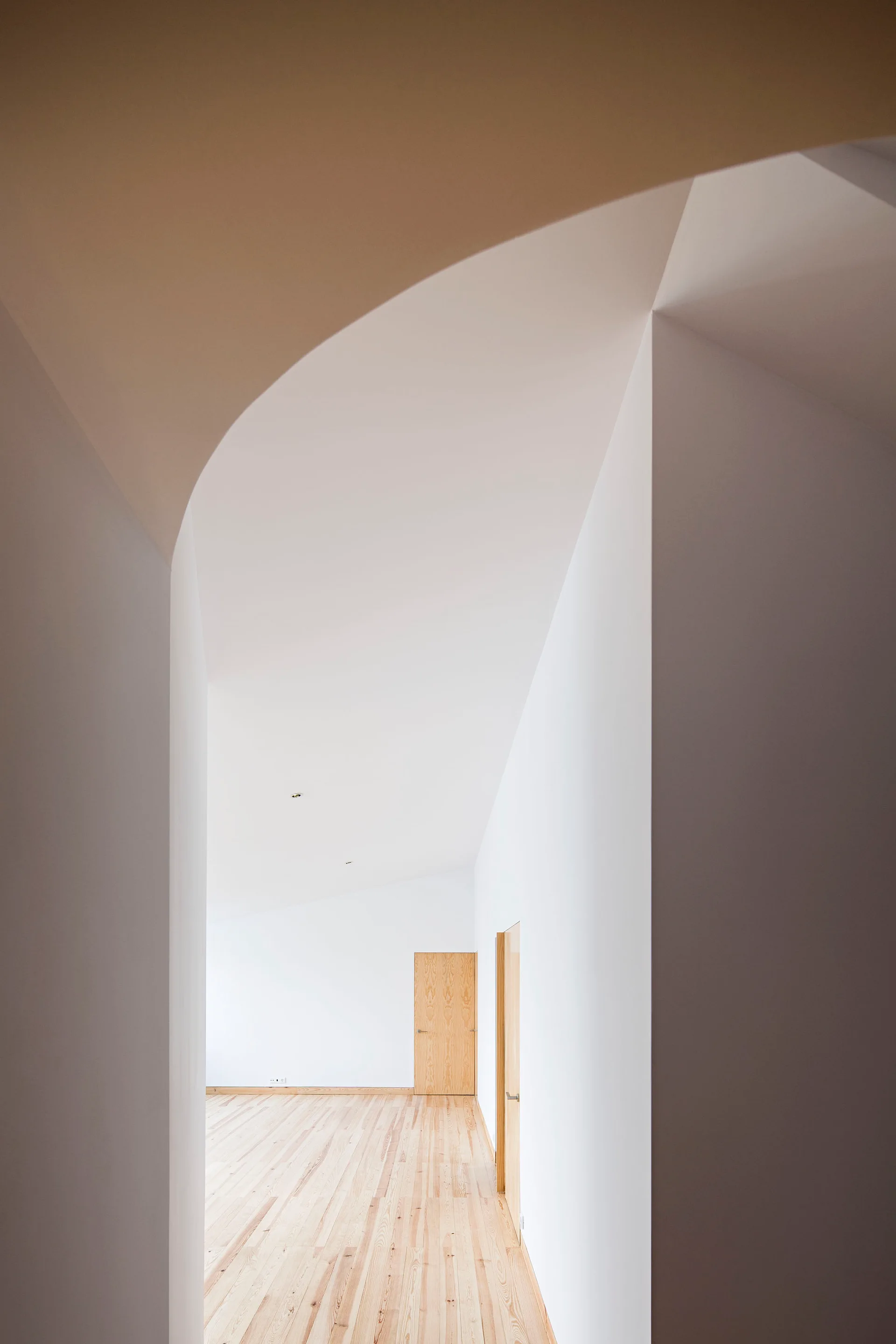A minimalist hallway viewed through a curved plaster archway, featuring smooth white walls, light wood flooring, and natural wood doors.