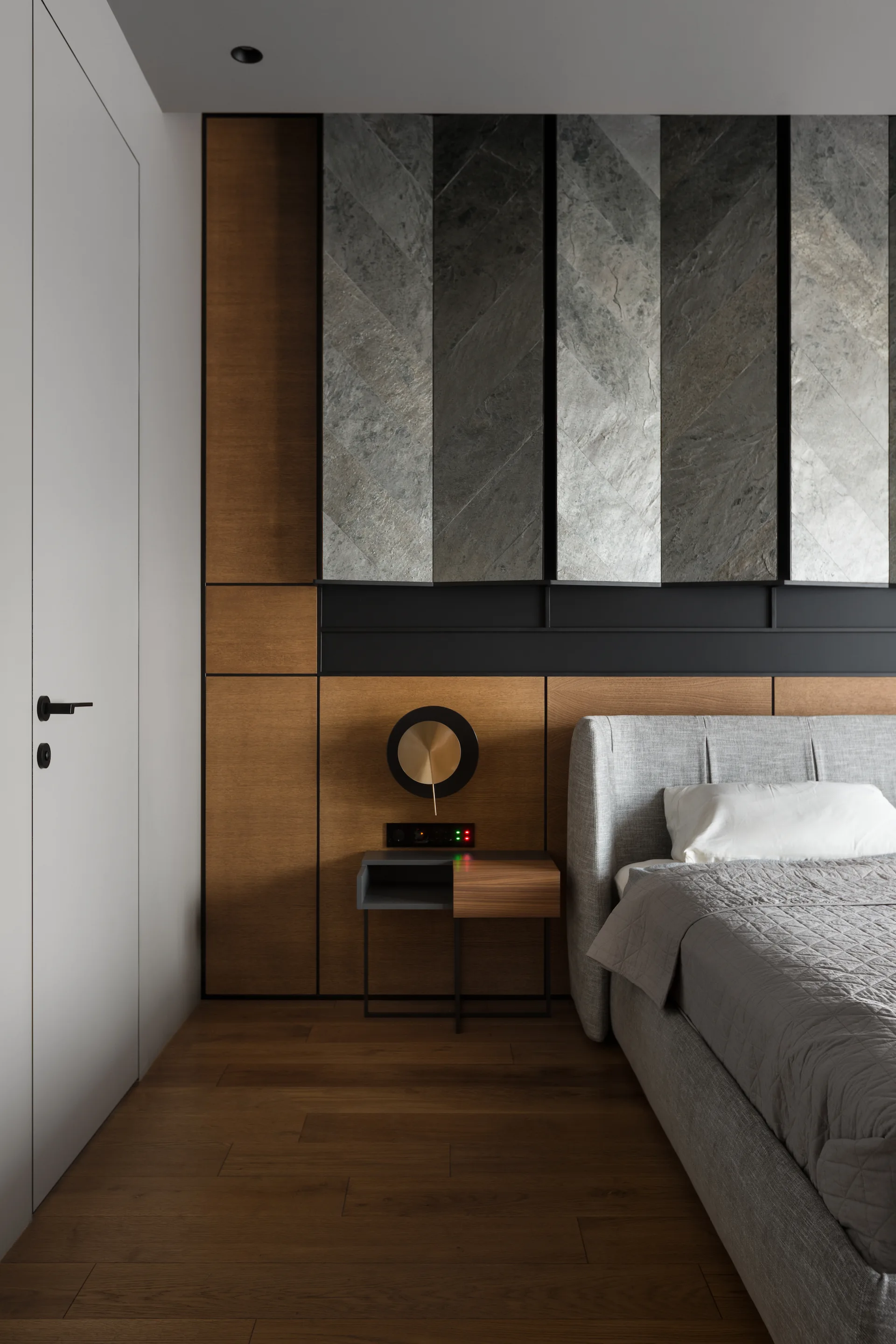 A modern bedroom features a grey upholstered bed next to a wood-paneled wall with herringbone stone tiles and a circular wall sconce.