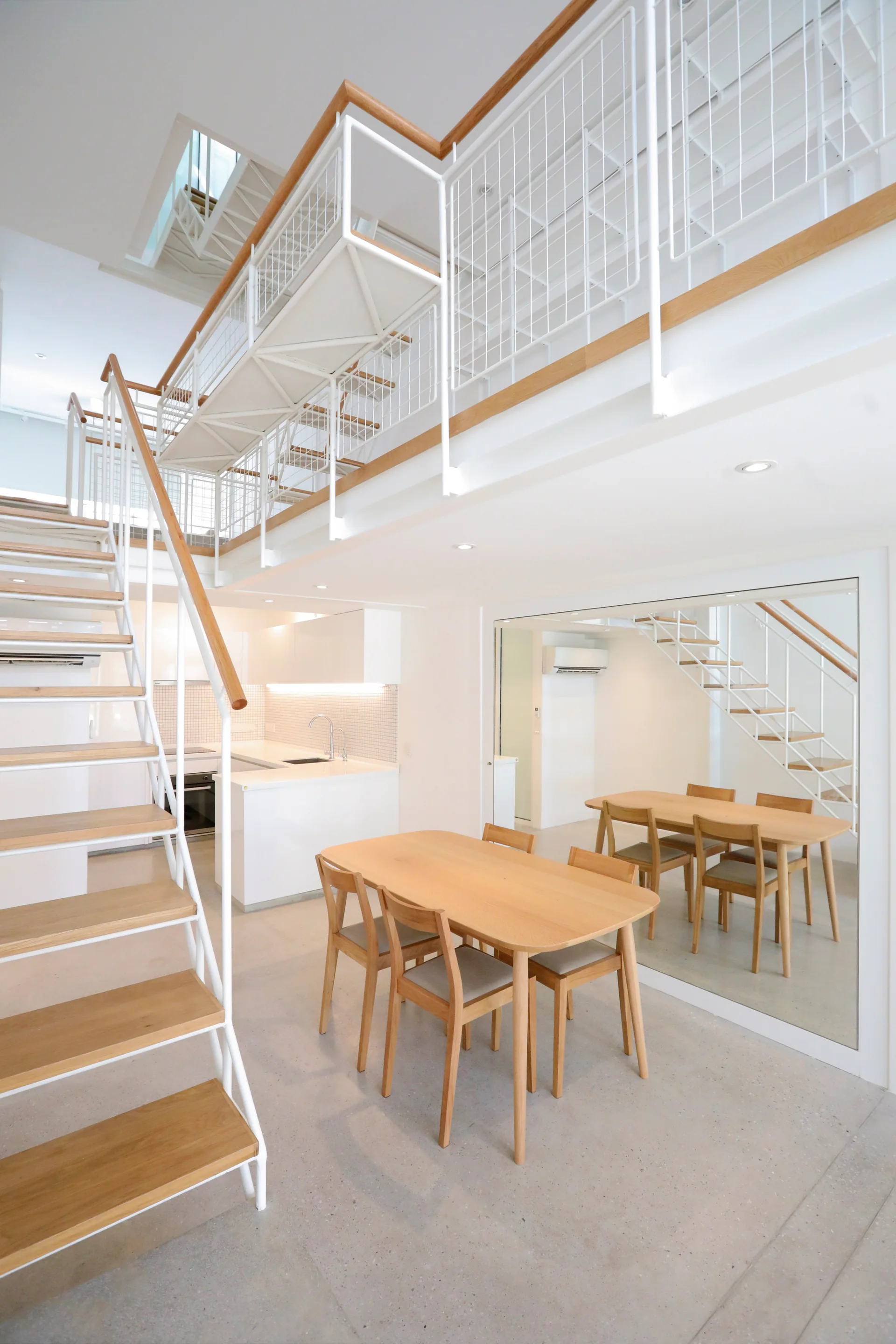 A minimalist dining room features a light wood table and chairs next to a white open-tread staircase and a large wall mirror.
