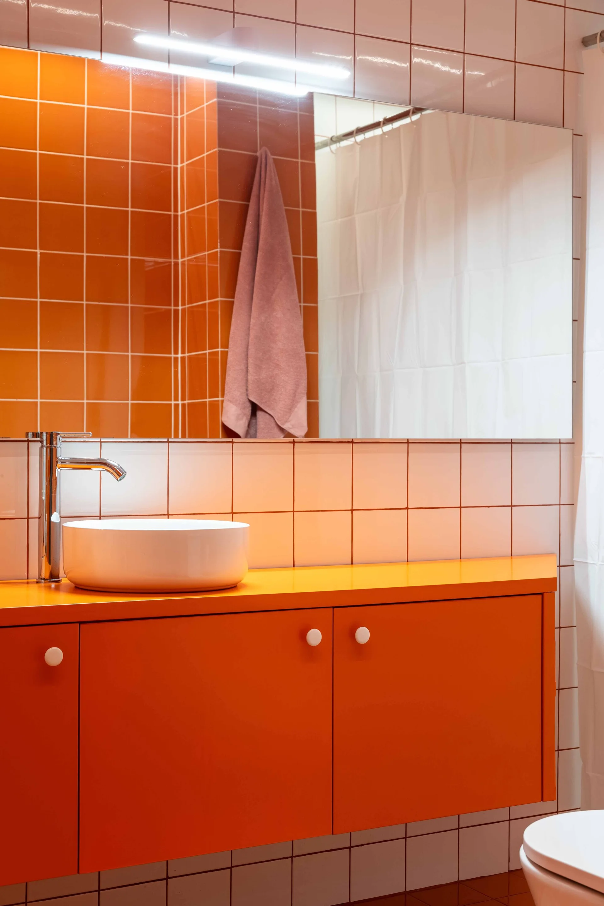 A modern bathroom featuring a bright orange floating vanity, a white vessel sink with a chrome faucet, and glossy orange square-tiled walls.