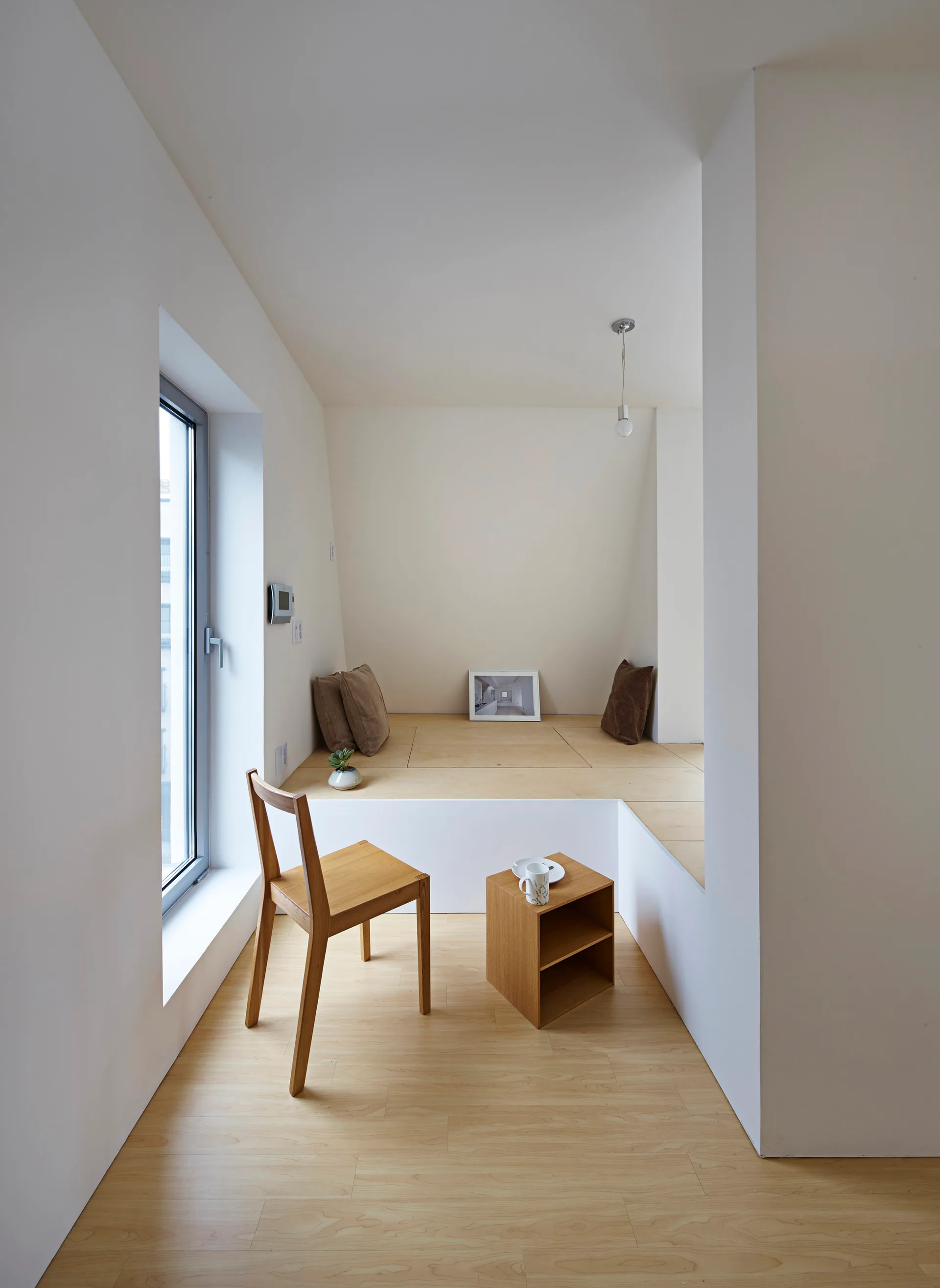 A minimalist room features a raised wooden platform with brown cushions, a simple wooden chair, and a small wooden side table next to a window.