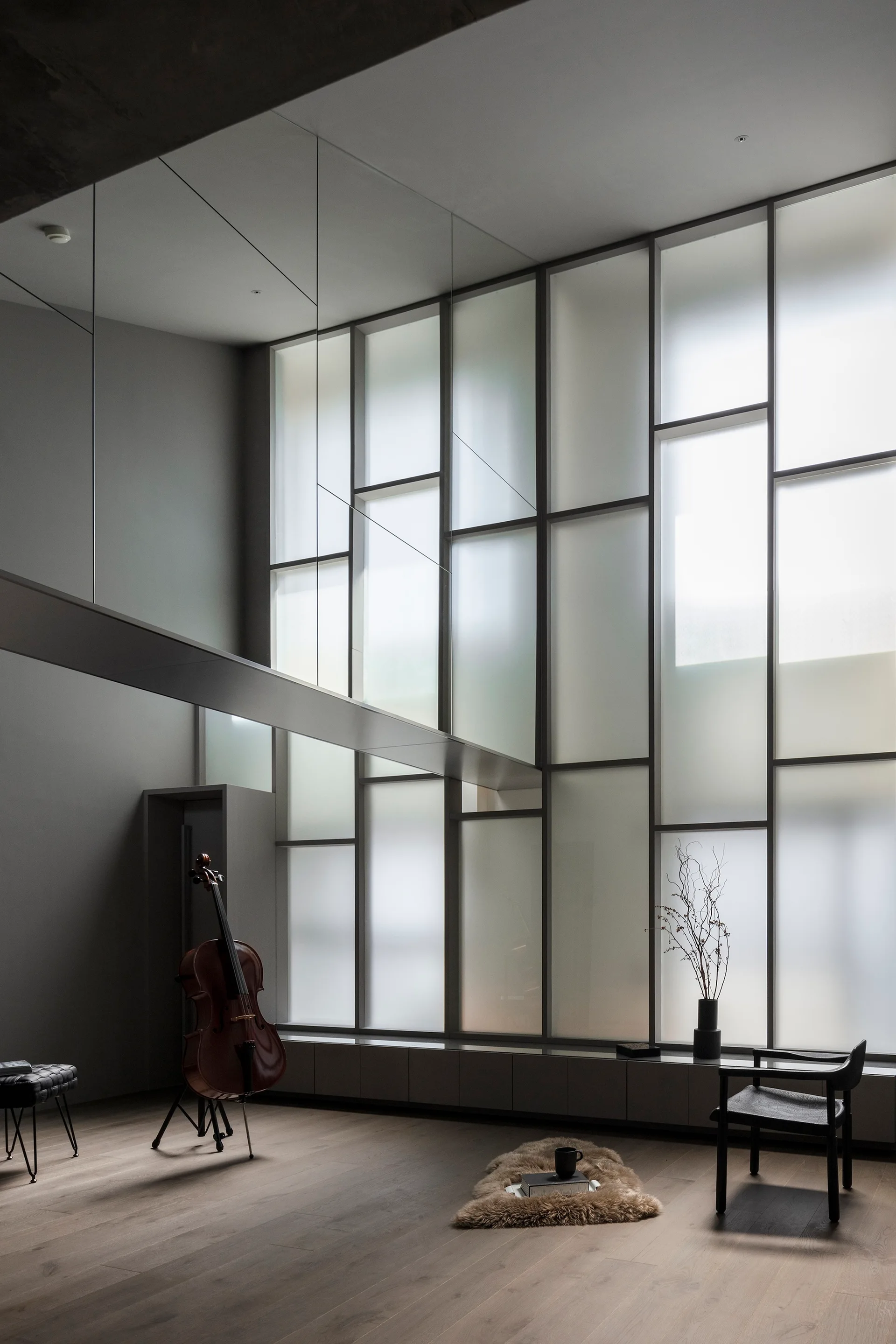 A minimalist grey room features a tall frosted glass grid window wall, natural wood flooring, a cello on a stand, and a black accent chair.