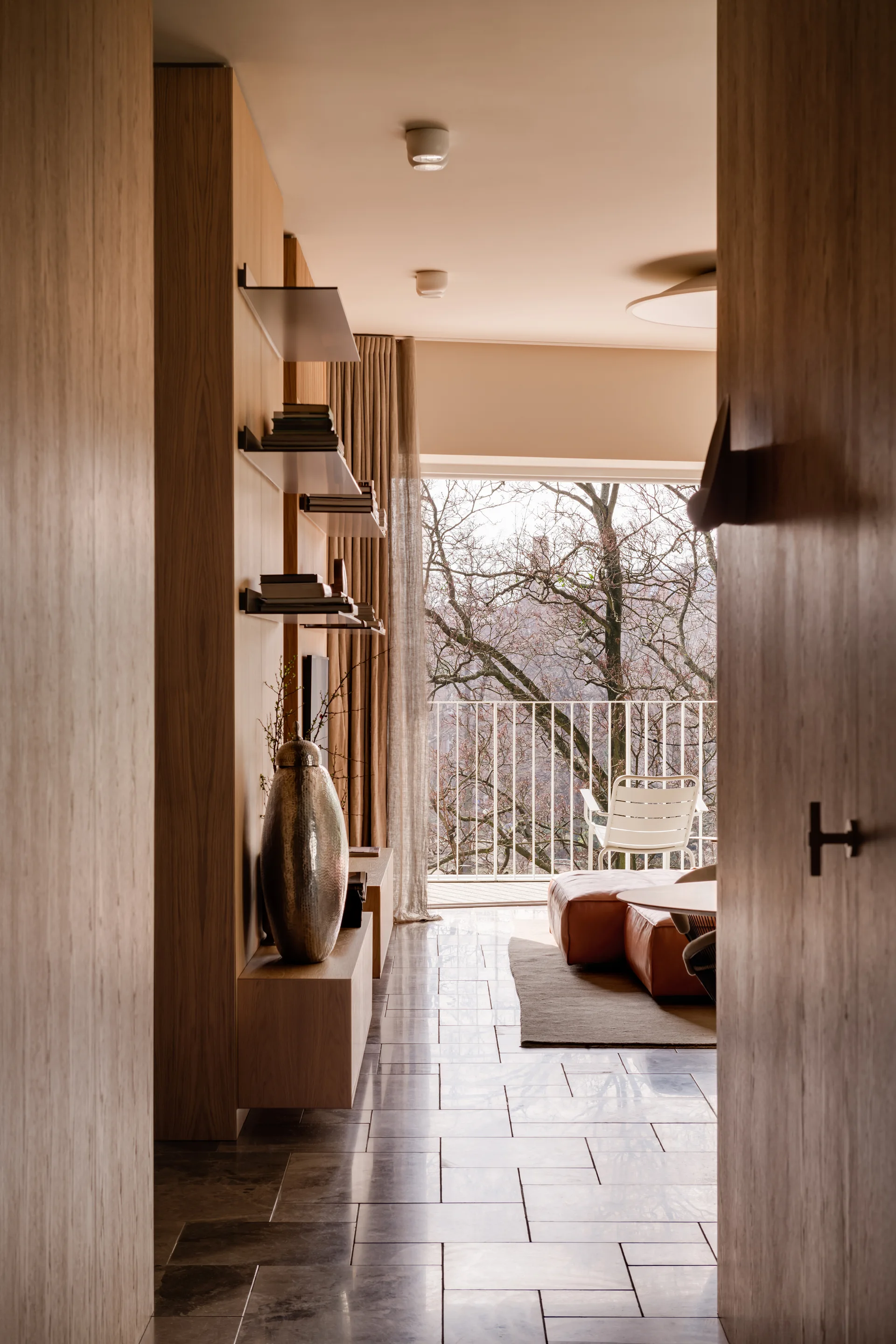 A modern living room viewed from a hallway, featuring wood panel walls with built-in shelves, a stone tile floor, and a balcony window.