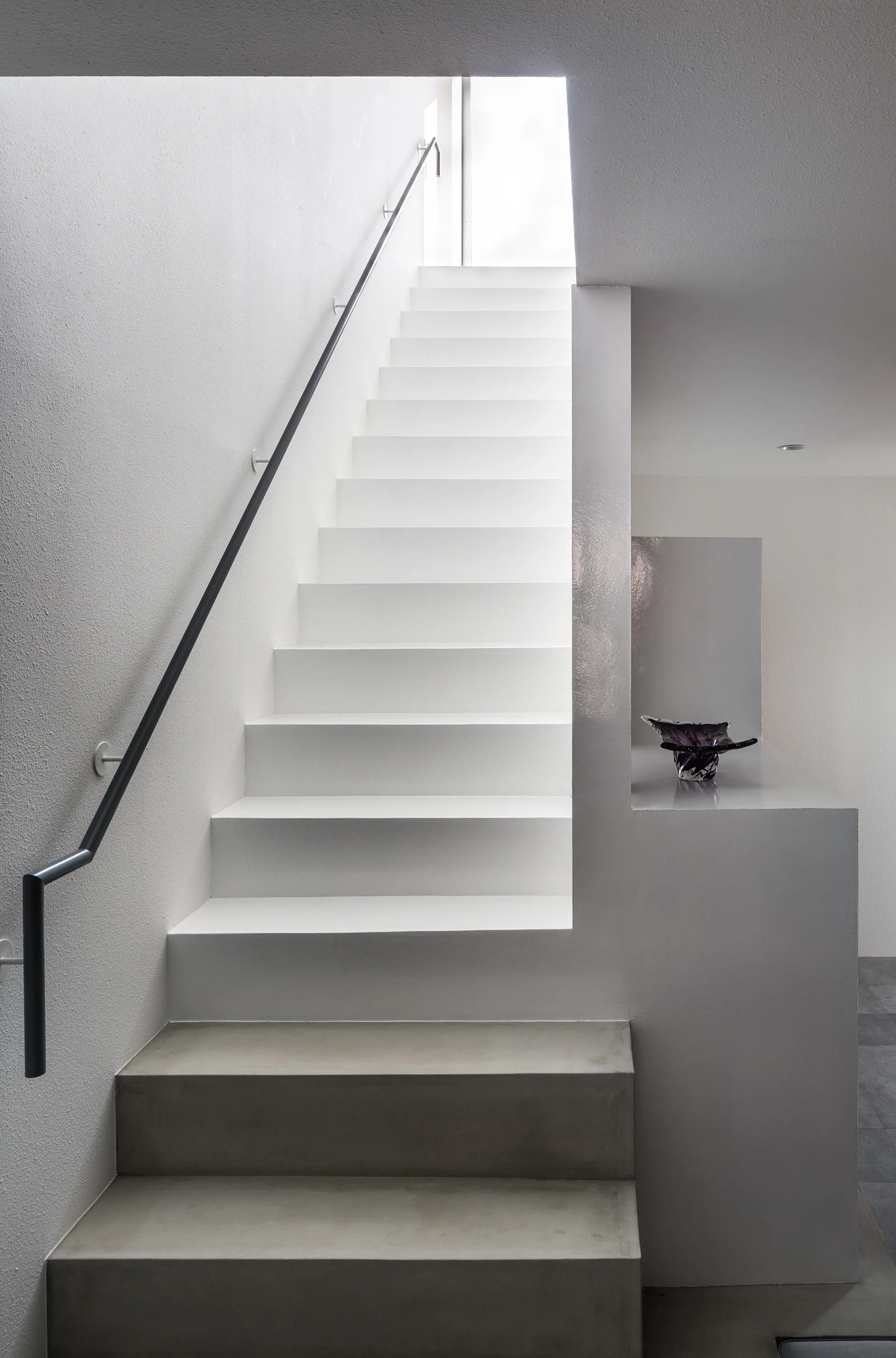 A minimalist white staircase features grey concrete base steps and a thin black metal handrail under bright natural overhead lighting.