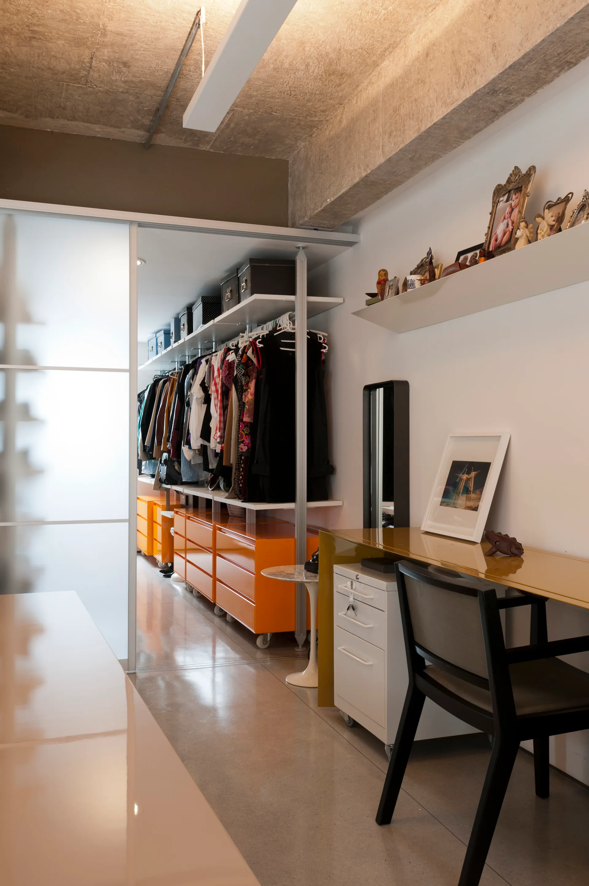 A modern walk-in closet featuring bright orange drawer units and clothing racks adjacent to a desk area beneath an exposed concrete ceiling.
