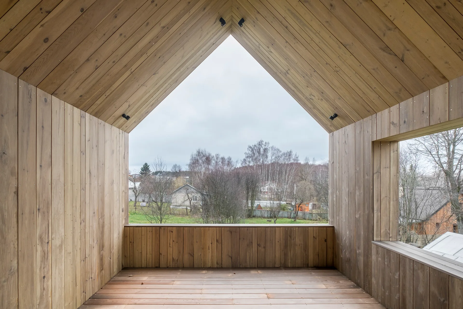 An elevated wooden balcony wrapped in natural timber planks features a gabled opening looking out onto a rural landscape under an overcast sky.