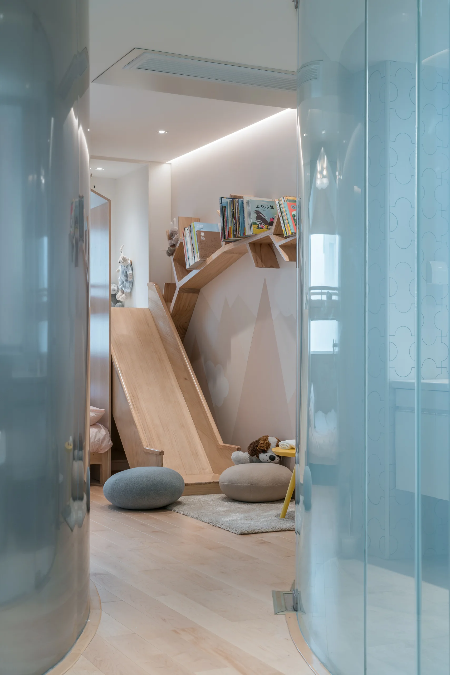A children's room featuring a custom wooden indoor slide, a tree-shaped bookshelf, mountain wall decor, and curved light blue glass panels.