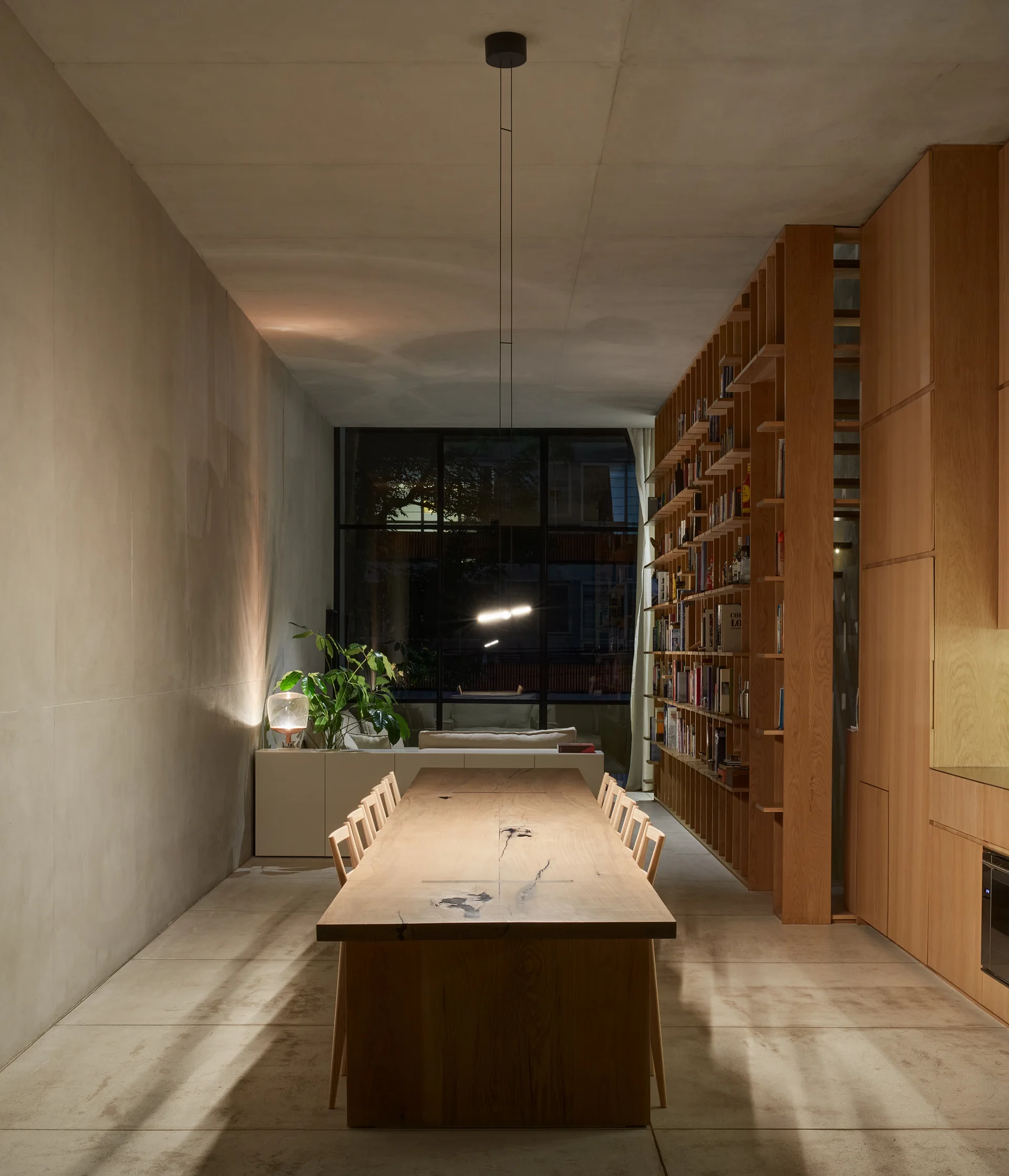 A long wooden dining table and chairs sit between a concrete wall and a floor-to-ceiling wooden bookcase in a minimalist room at night.