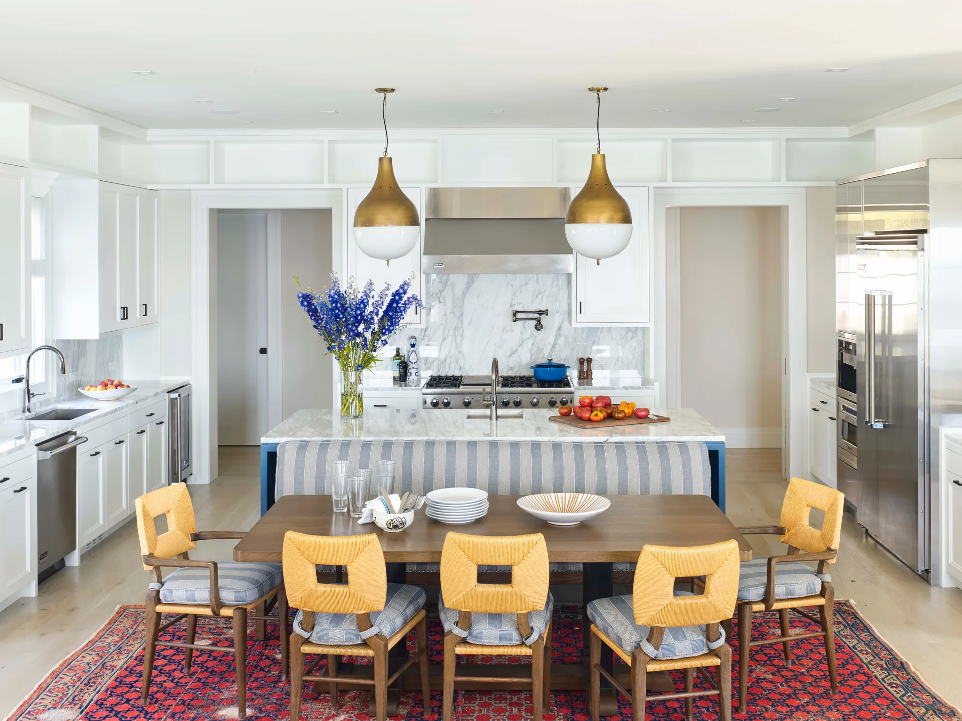 A bright kitchen featuring a blue island with an integrated striped banquette, a wooden dining table, yellow woven chairs, and brass pendant lights.