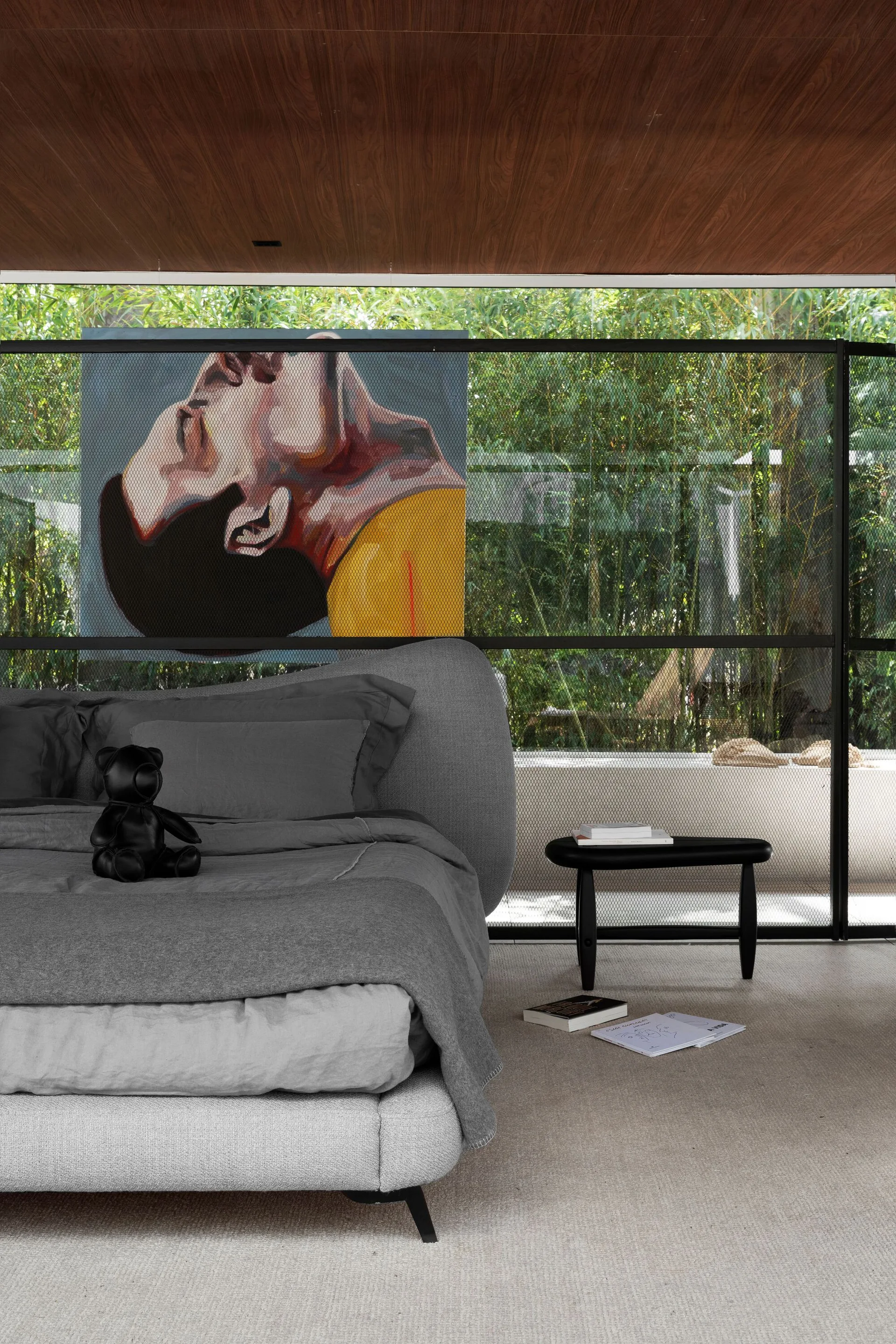 A modern bedroom features a grey upholstered bed in front of a black metal mesh screen displaying an art piece under a wood plank ceiling.